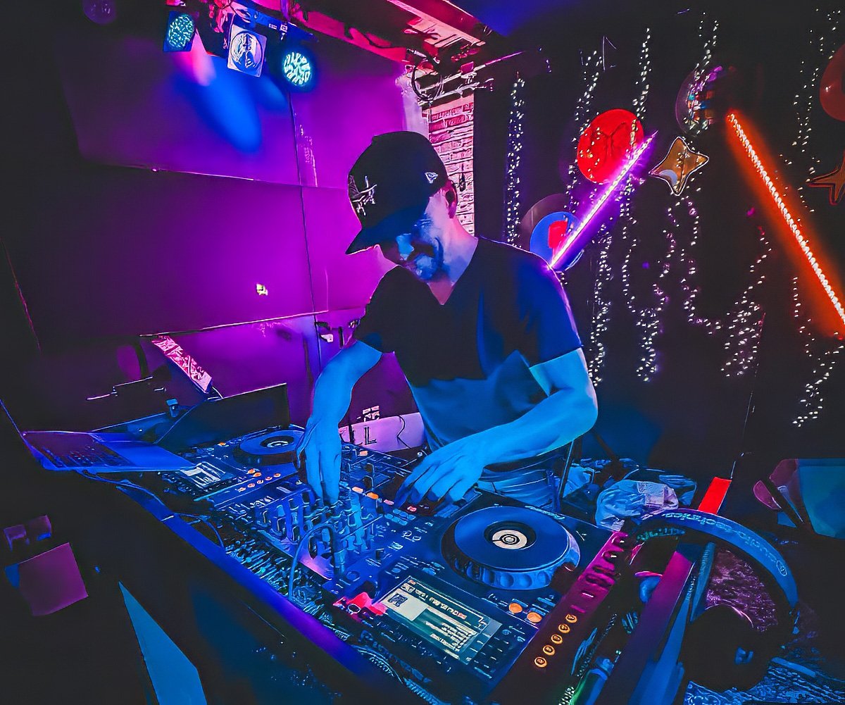 A DJ wearing a black t-shirt and a baseball cap performing at a nightclub with colorful lights and decorative balloons in the background.