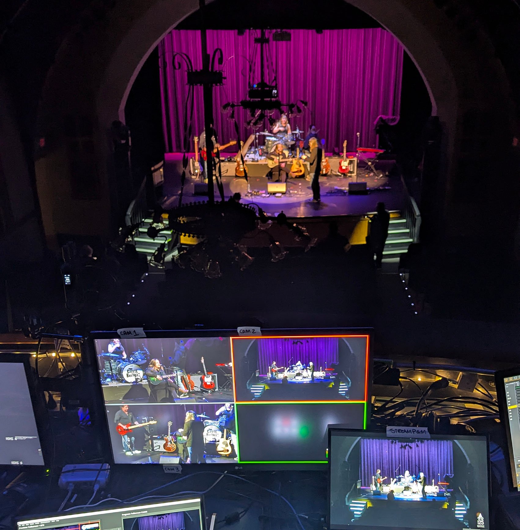 Image of multiple monitors showing footage of a live musical performance on stage with a purple curtain background. The stage features musicians playing guitars, keyboards, and drums with a singer performing in the center. Audience silhouettes are visible in the foreground.