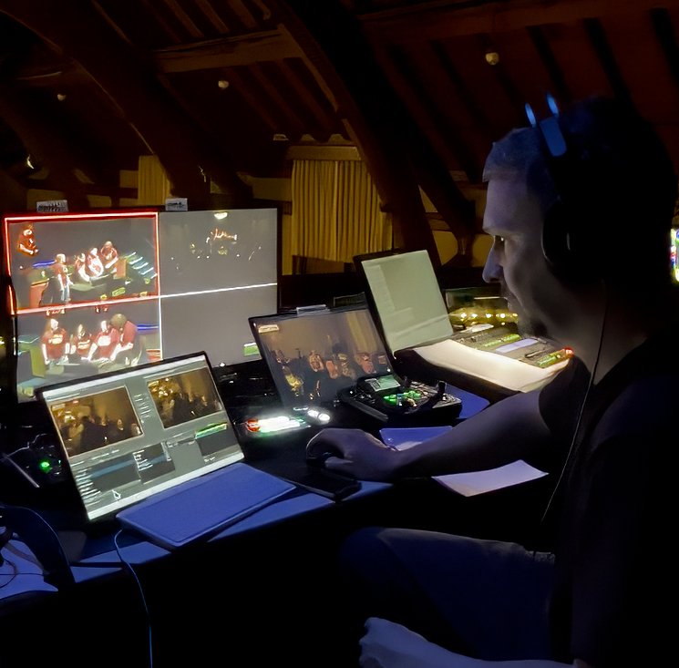 A person operating multiple monitors and audio equipment at a live event or broadcast control room, with a young man wearing headphones.