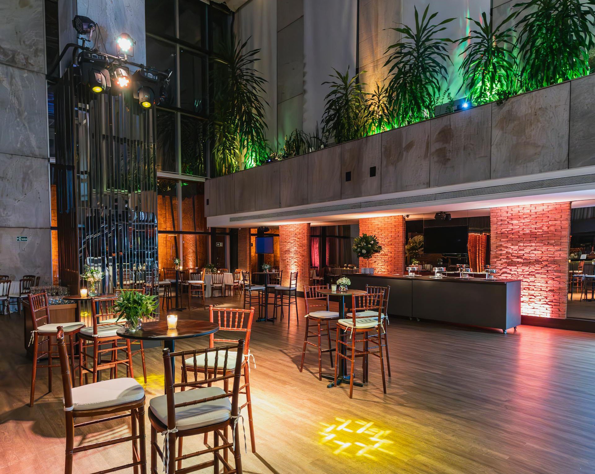 An event space with round tables and chairs, decorated with flower centerpieces and candles, indoors with a bar area, exposed brick wall, and colored lighting, suitable for a celebration or reception.