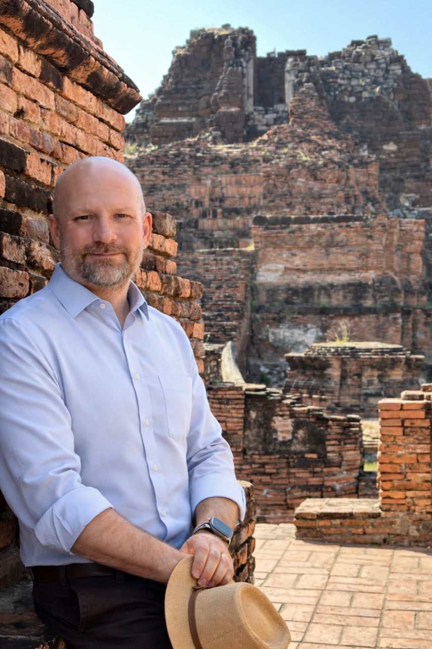 A man with a beard dressed in a light blue shirt holding a tan hat, standing next to a brick wall with ancient ruins and a stepped pyramid structure in the background.