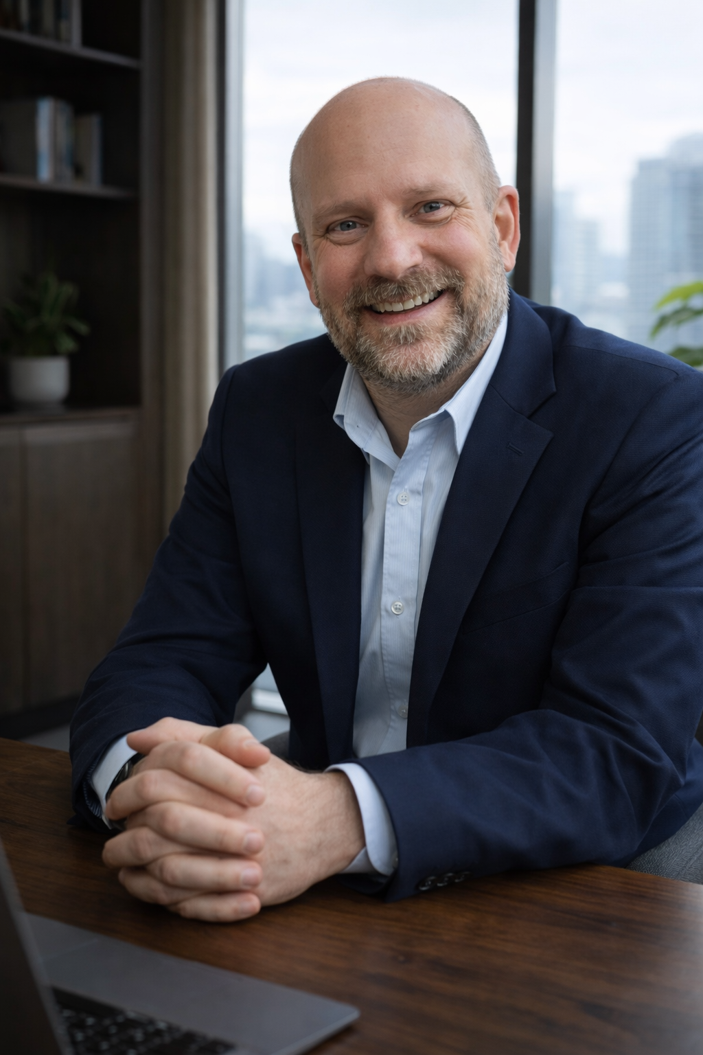 A smiling middle-aged man with a beard and bald head, wearing a navy blazer and light blue dress shirt, sitting at a wooden desk in an office with large windows showing city buildings in the background.