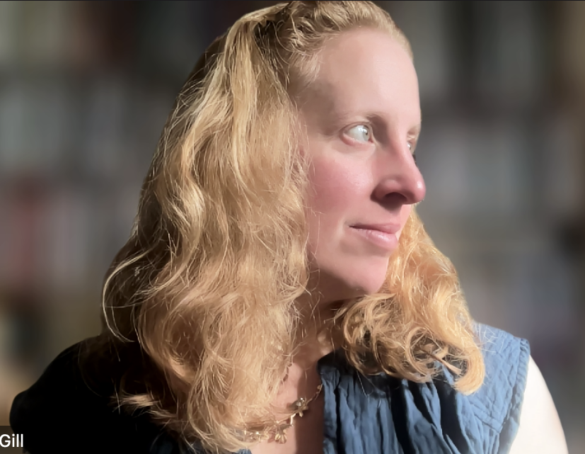 A photo of Ailithir O. McGill looking to her right, with her strawberry-blonde hair hanging in waves around her face, wearing a blue vest over a white top, in front of a blurred background.