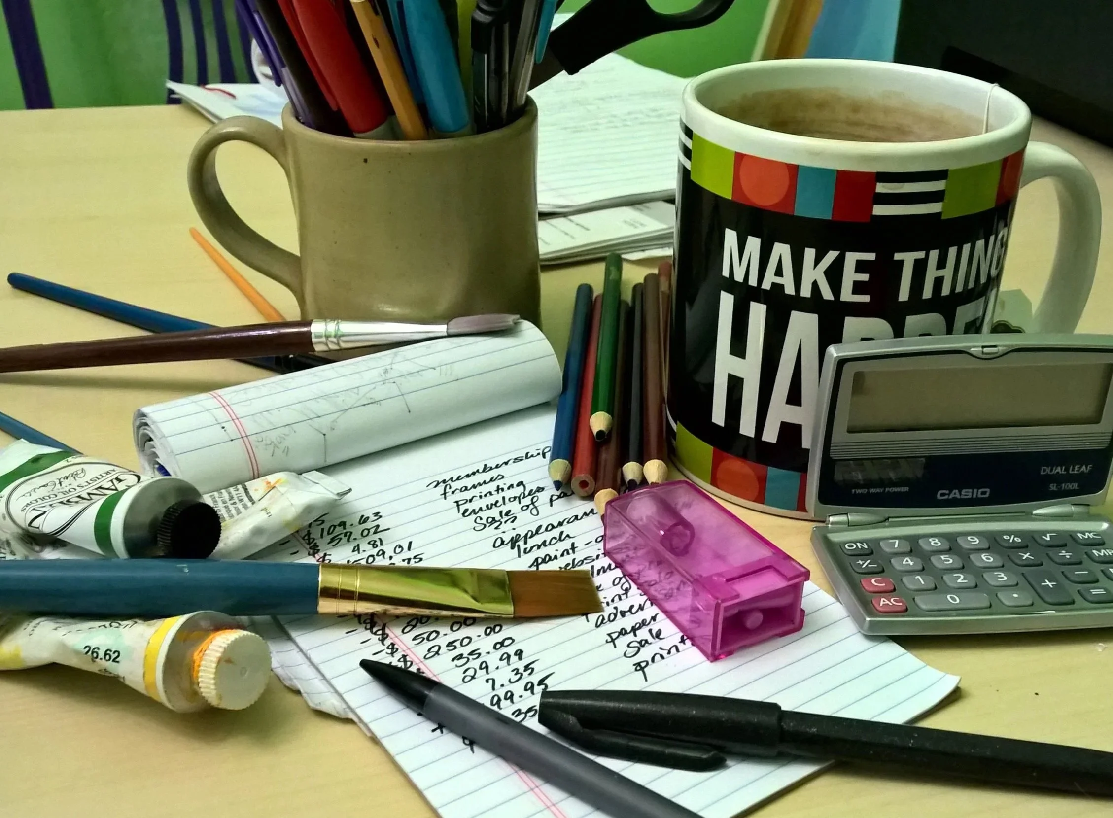 Messy desk with notes, pens, pencils, a calculator, a pink pencil sharpener, coffee mug with 'Make Things Happen' text, and a bottle of glue.