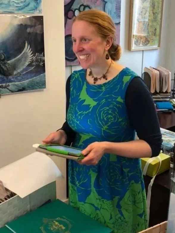 A photo of Ailithir O. McGill, wearing a blue and green patterned dress and a necklace, smiling and selling copies of her book.
