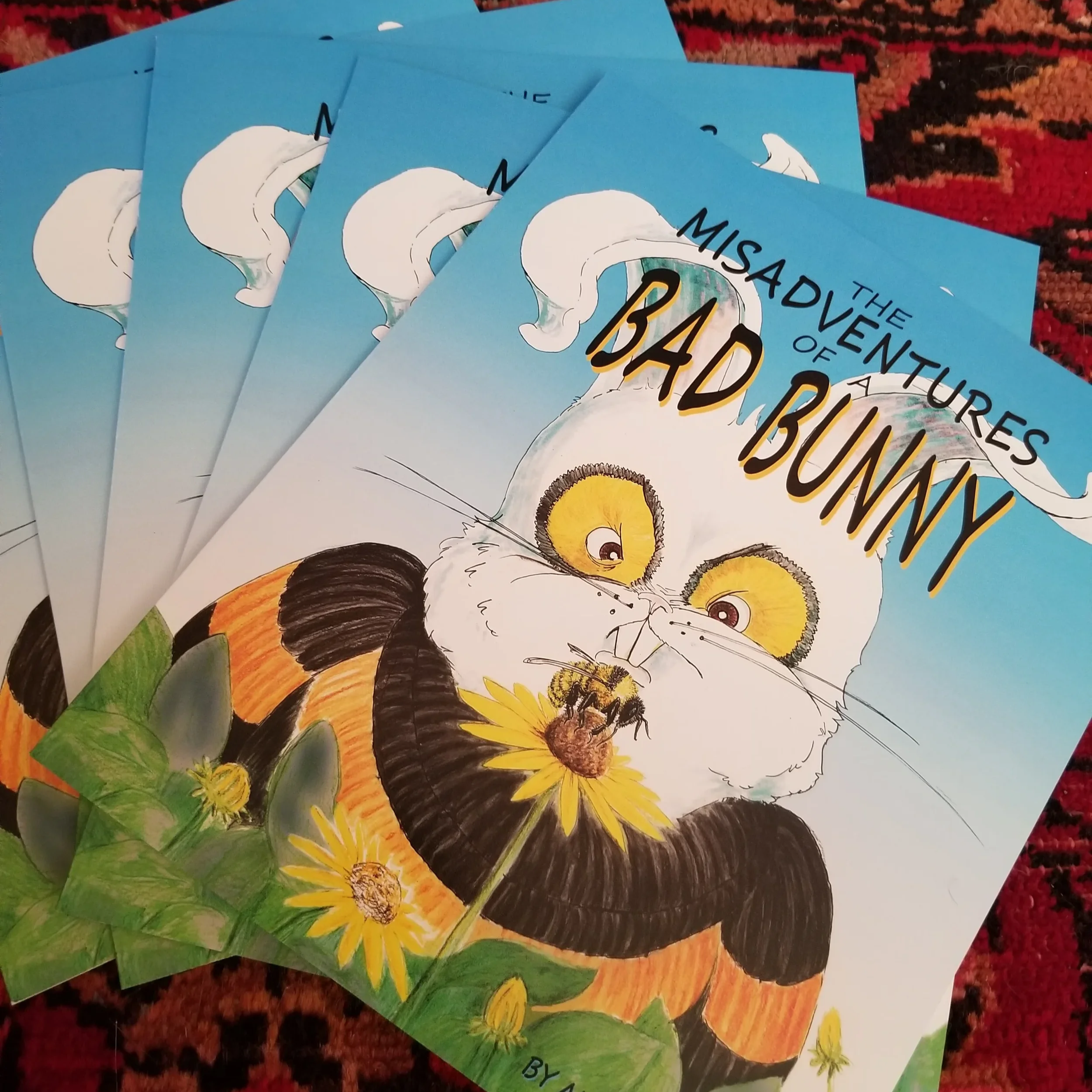 Multiple copies of a book titled "The Misadventures of a Bad Bunny" with a colorful illustration of a bunny glaring at a bumblebee on the cover, featuring yellow flowers and green foliage, set on a patterned surface.
