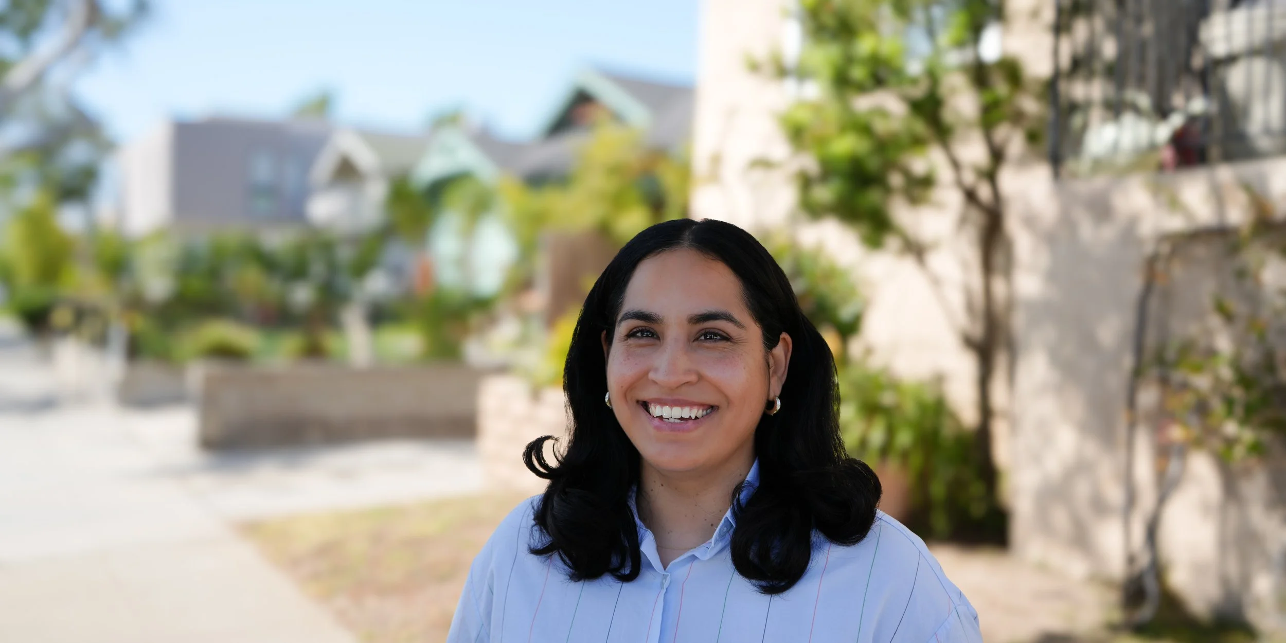 A woman with dark hair smiling outdoors with houses, trees, and a blue sky in the background.