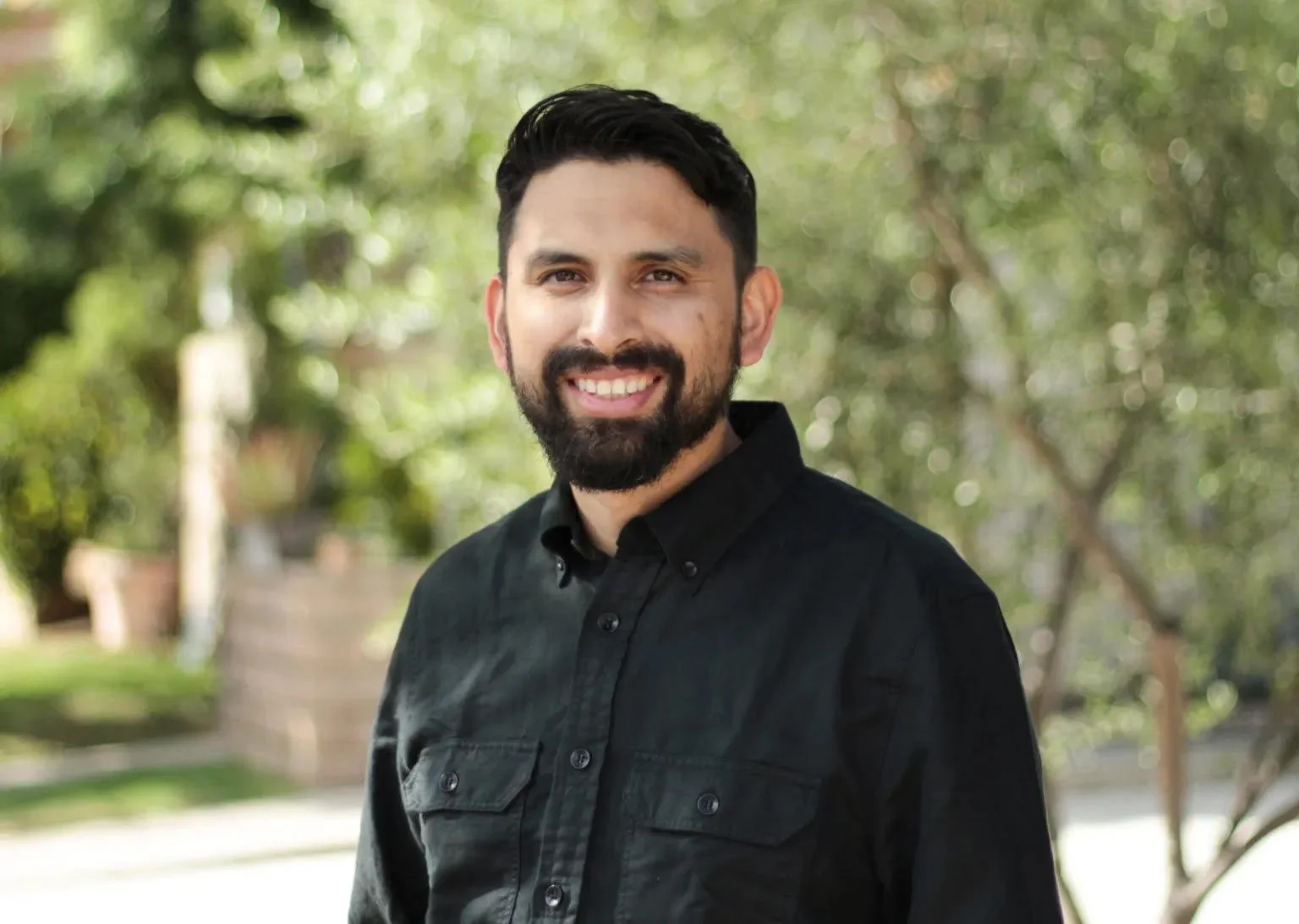 A smiling man with dark hair and a beard wearing a black button-up shirt standing outdoors with a background of trees and greenery.