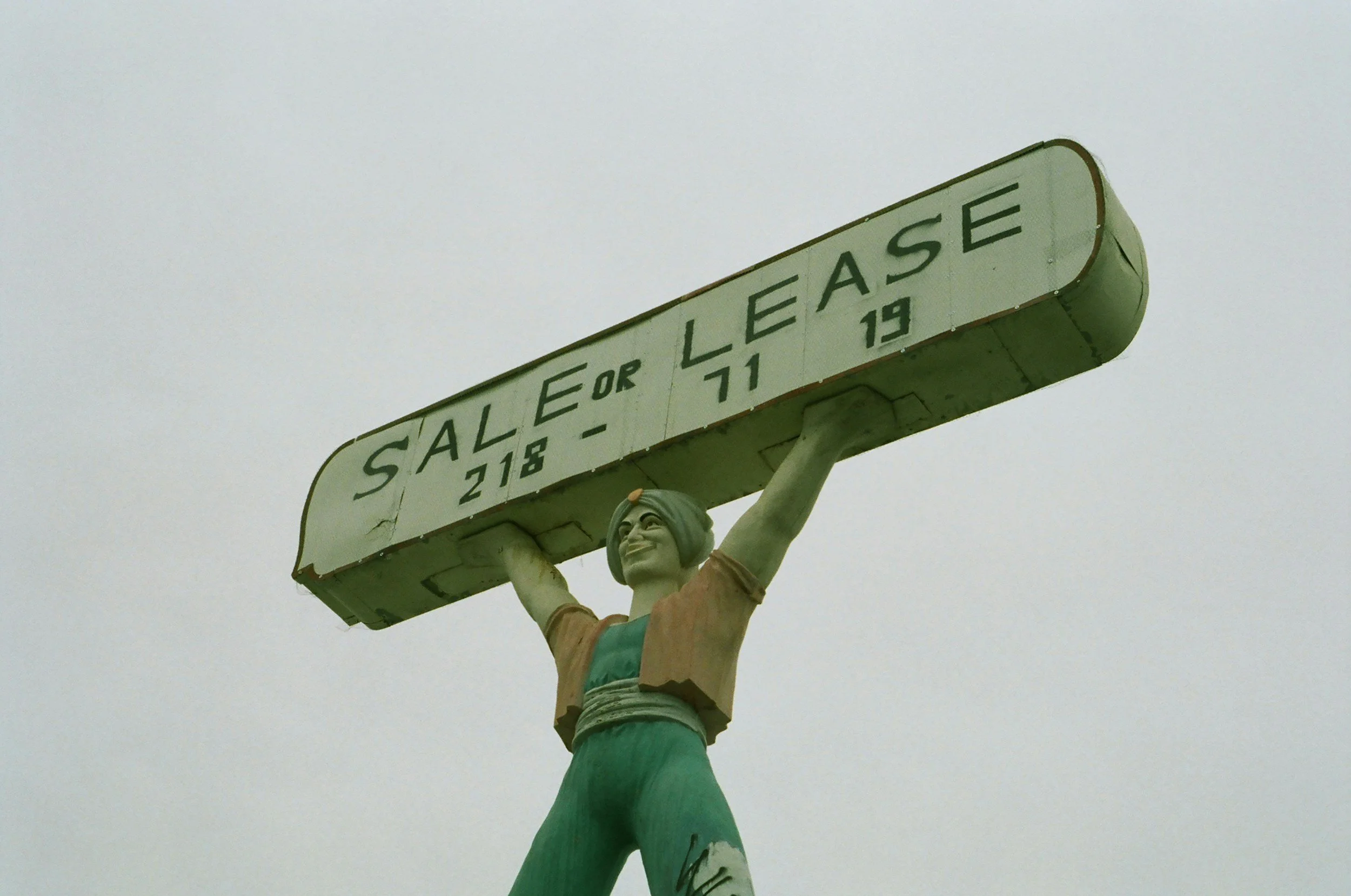 Statue of a woman holding a sign that reads 'Sale or Lease 218-71 19' against a cloudy sky.