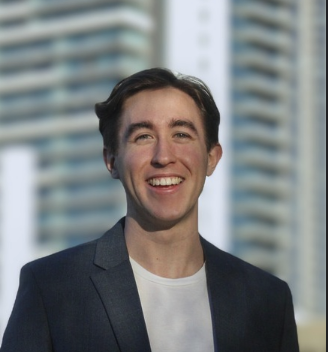A young man with dark hair smiling, wearing a dark blazer and white shirt, standing outdoors in front of tall modern buildings.