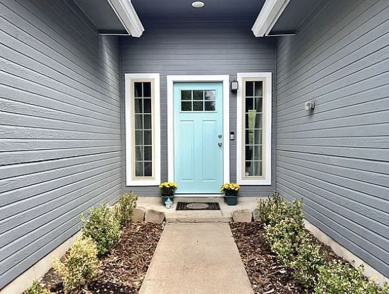 Front porch of a house with gray siding and a light blue door, flanked by windows and potted flowers, with a concrete walkway leading up to it.