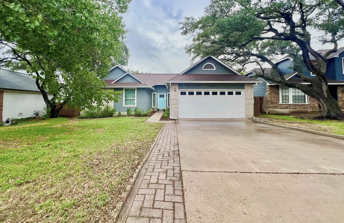 Front view of a blue house with a white garage door, surrounded by trees and a lawn, with a brick-paved pathway leading to the front door.