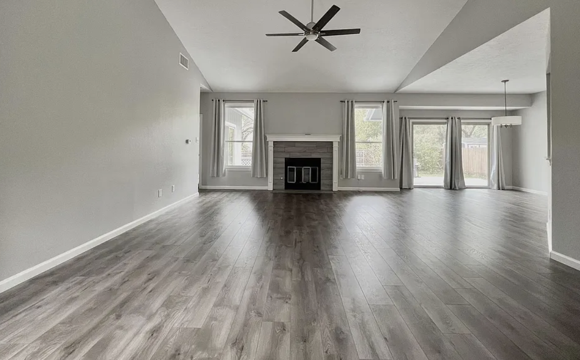 Empty living room with wood flooring, a central fireplace, light gray walls, three windows with curtains, and a ceiling fan.