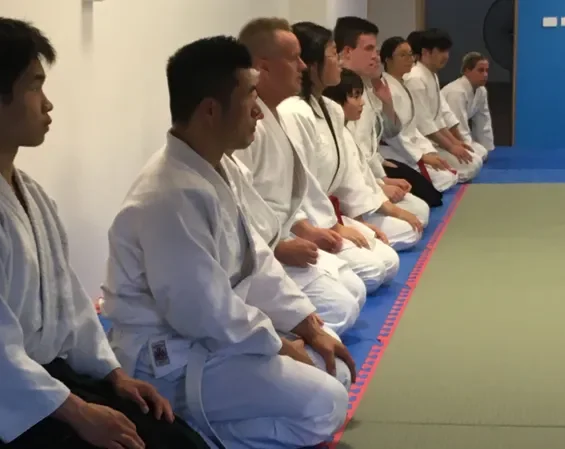 A group of martial artists sitting in a line on a mat in a dojo, wearing traditional gi uniforms.