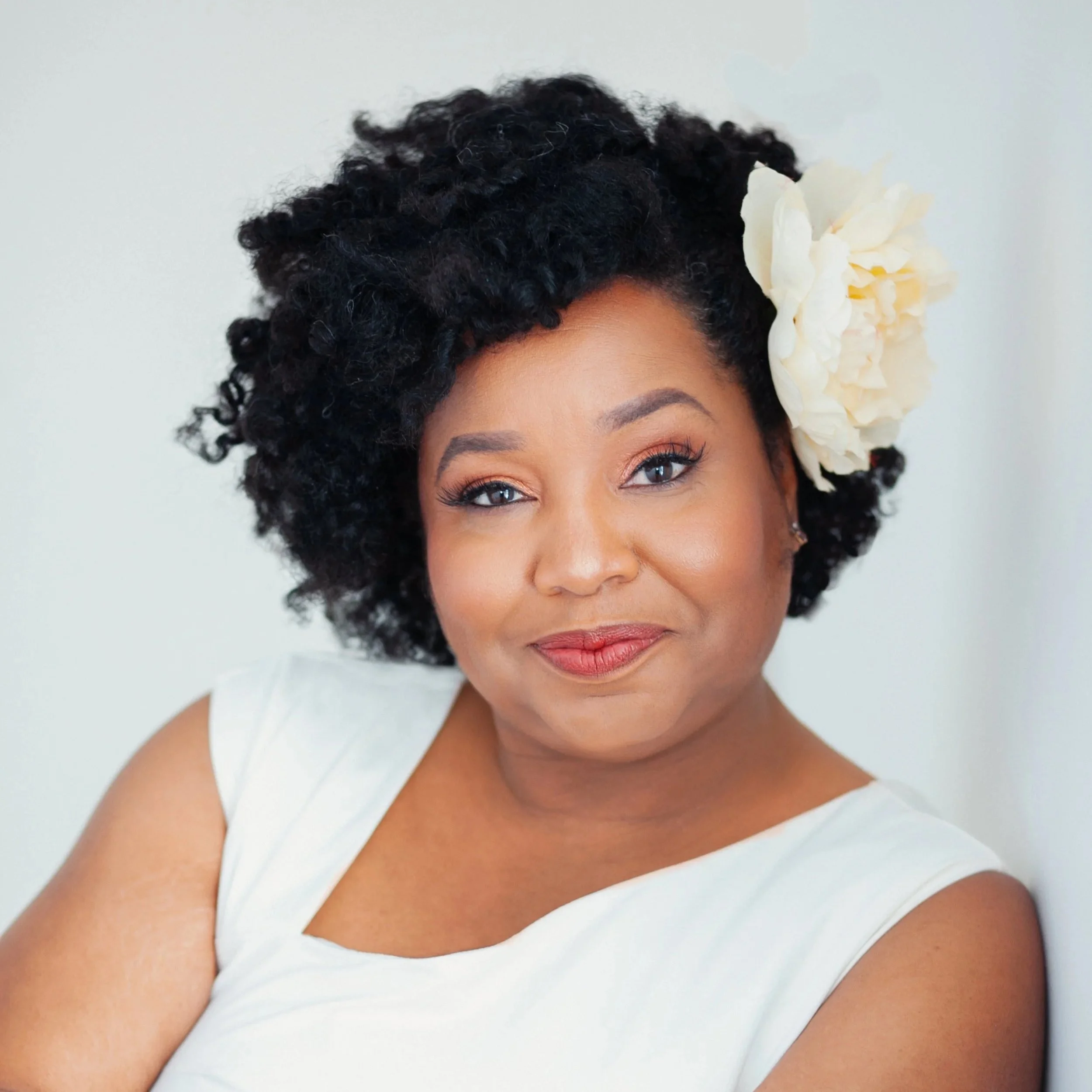 Portrait of a woman with short curly black hair and a large white flower in her hair, wearing a white sleeveless top, smiling softly against a plain background.