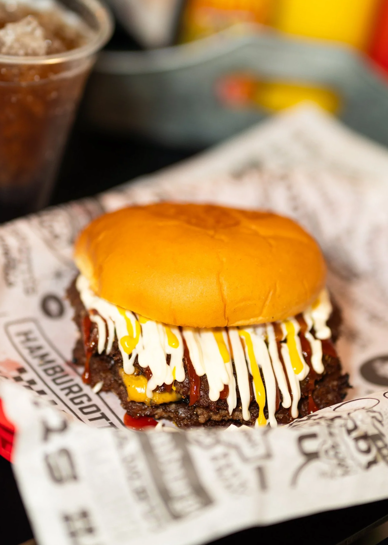 Close-up of a burger with a glossy bun, topped with mustard and mayonnaise, with a beef patty and ketchup visible underneath, served on branded paper.
