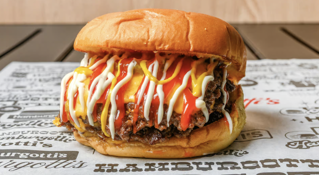Close-up of a cheeseburger with beef patty, melted cheese, ketchup, mustard, and mayonnaise in a bun, on paper with restaurant branding.