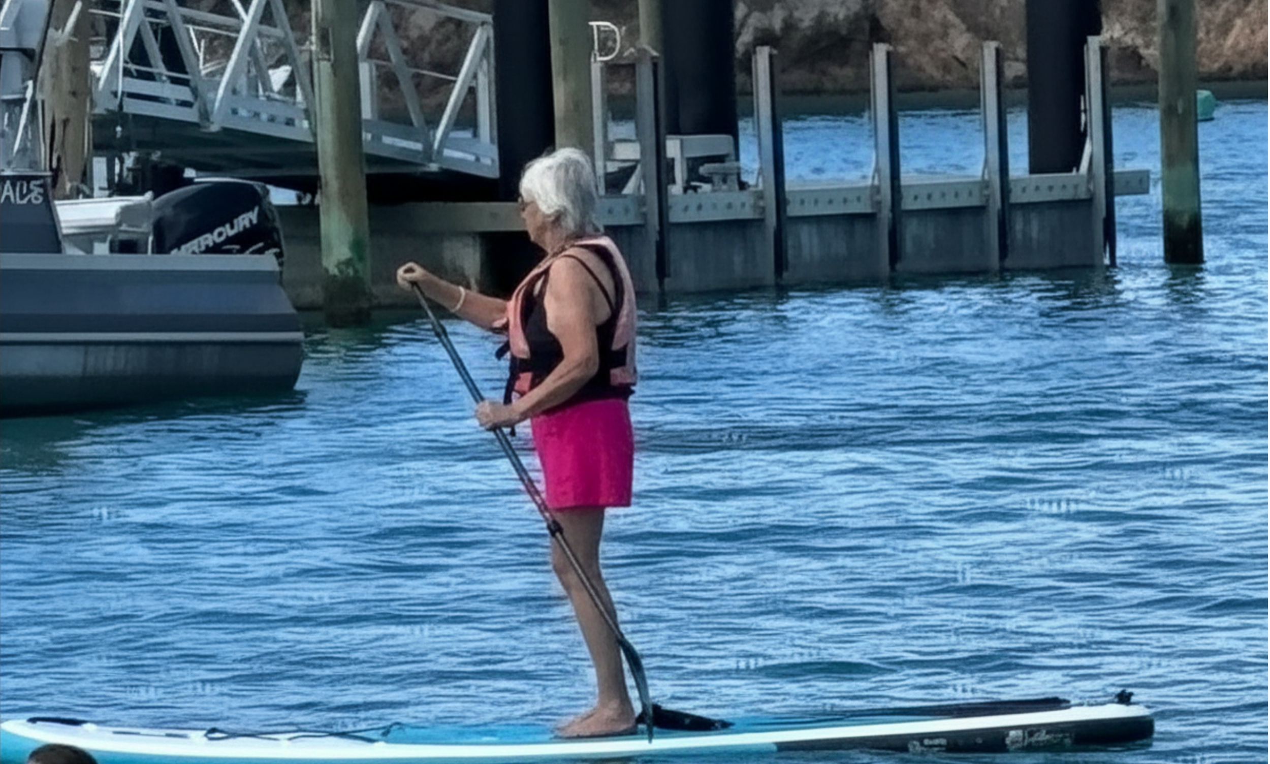 An elderly person stand-up paddleboarding on a calm body of water near a dock or pier. They are wearing a life jacket and pink shorts, holding a paddle, and have gray hair.
