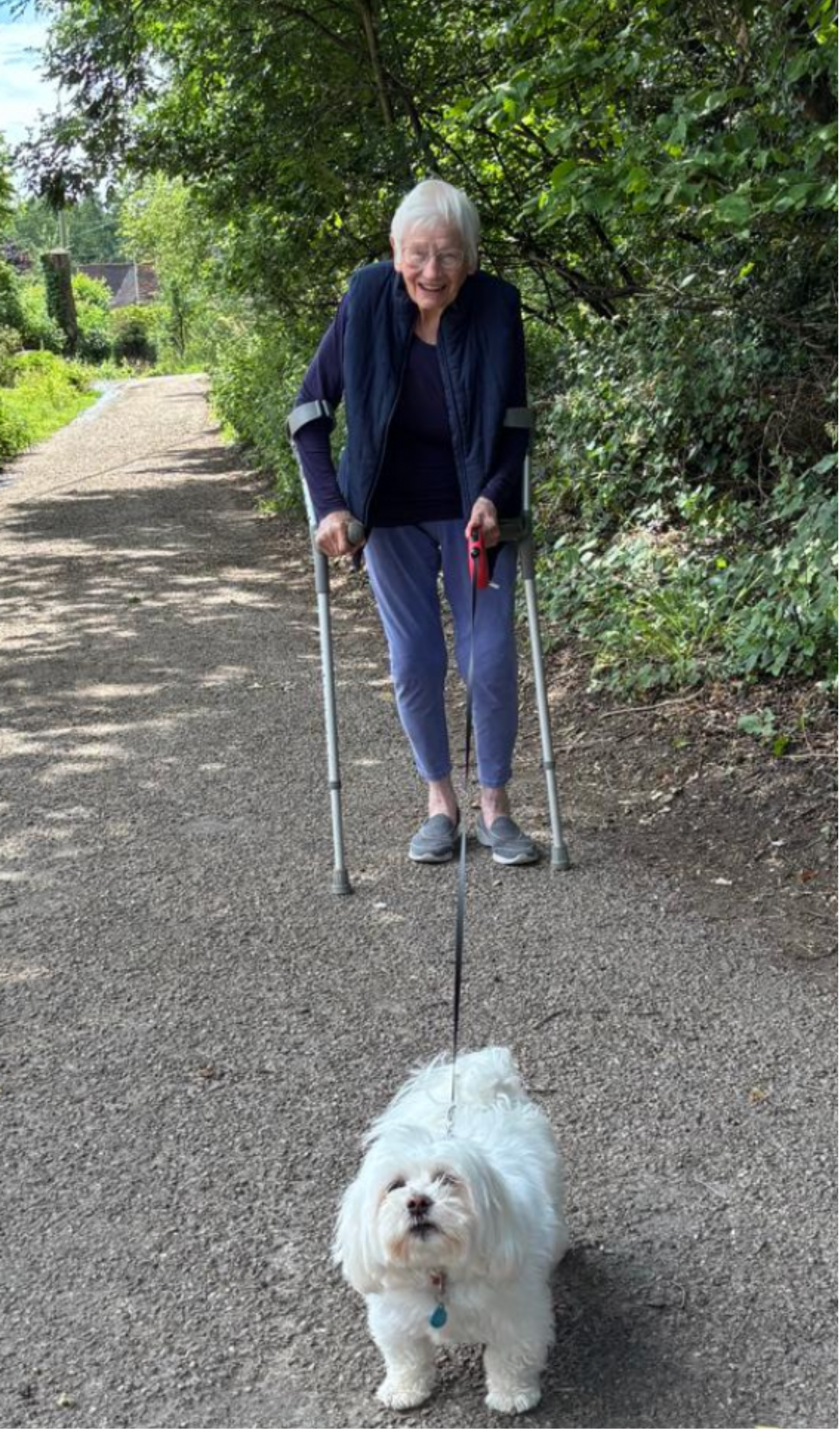 An elderly woman with white hair and glasses walking her small white fluffy dog on a dirt path surrounded by green trees and bushes.