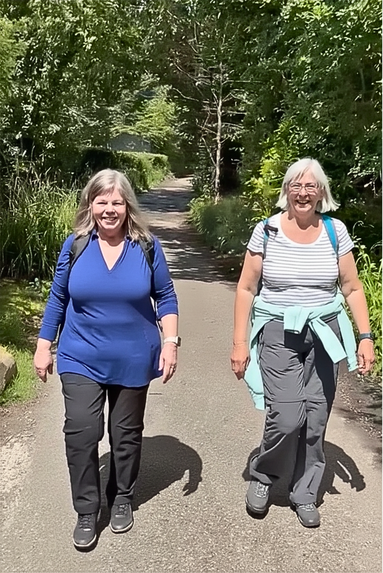 Two smiling women walking outdoors on a paved path surrounded by lush green trees and bushes. Both are dressed in casual hiking attire and wearing backpacks, enjoying a sunny day.