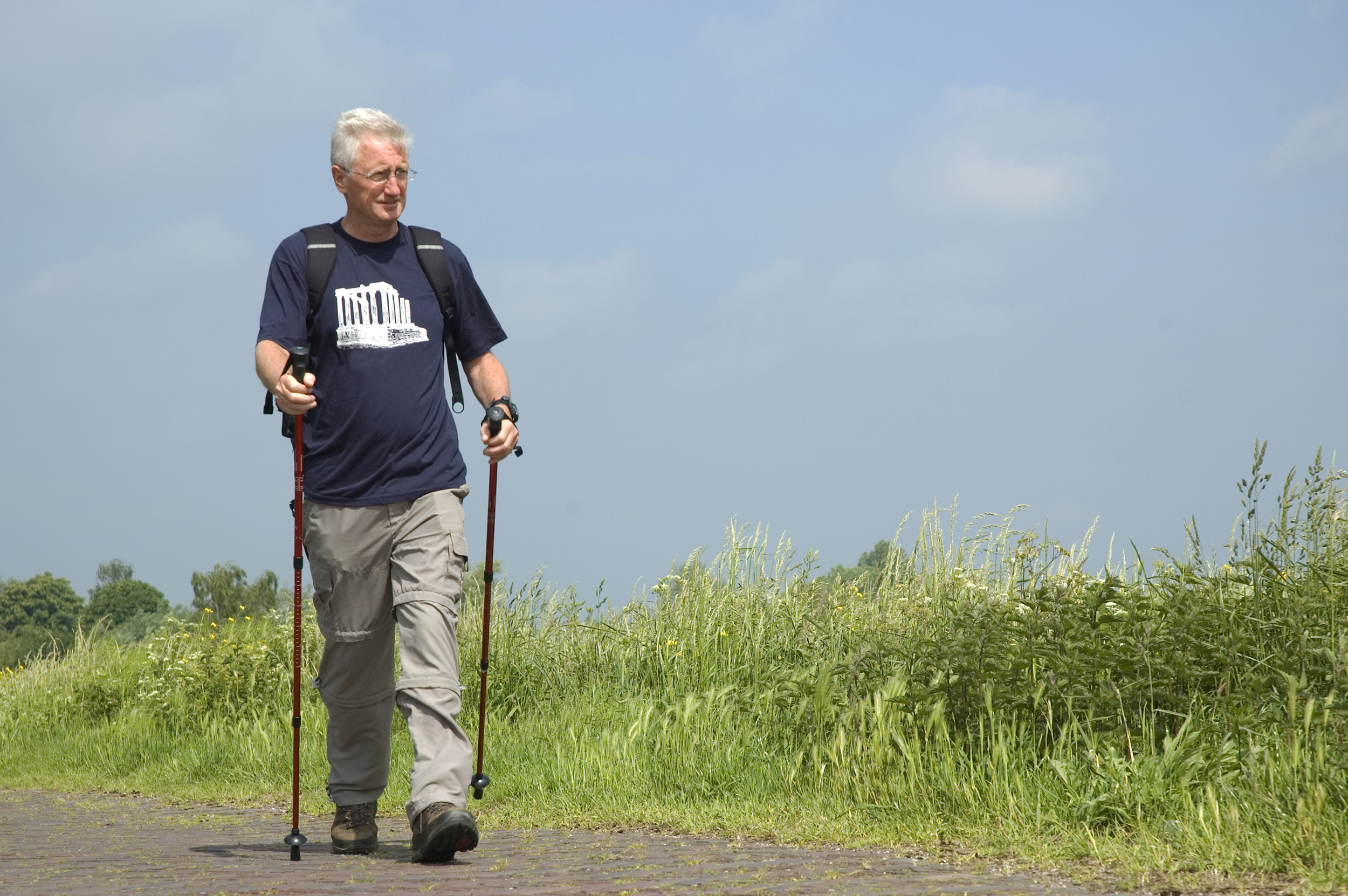 An older man with white hair, glasses, and wearing with a navy blue T-shirt with a building design, beige cargo pants, and hiking boots, is walking outdoors on a paved path with hiking poles. He has a backpack on and looks focused as he walks through