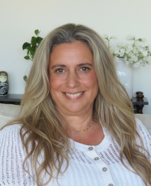 A smiling woman with long, wavy blonde hair sitting on a beige couch in a room with white walls, decorated with books, green plants, and white flowers in the background.