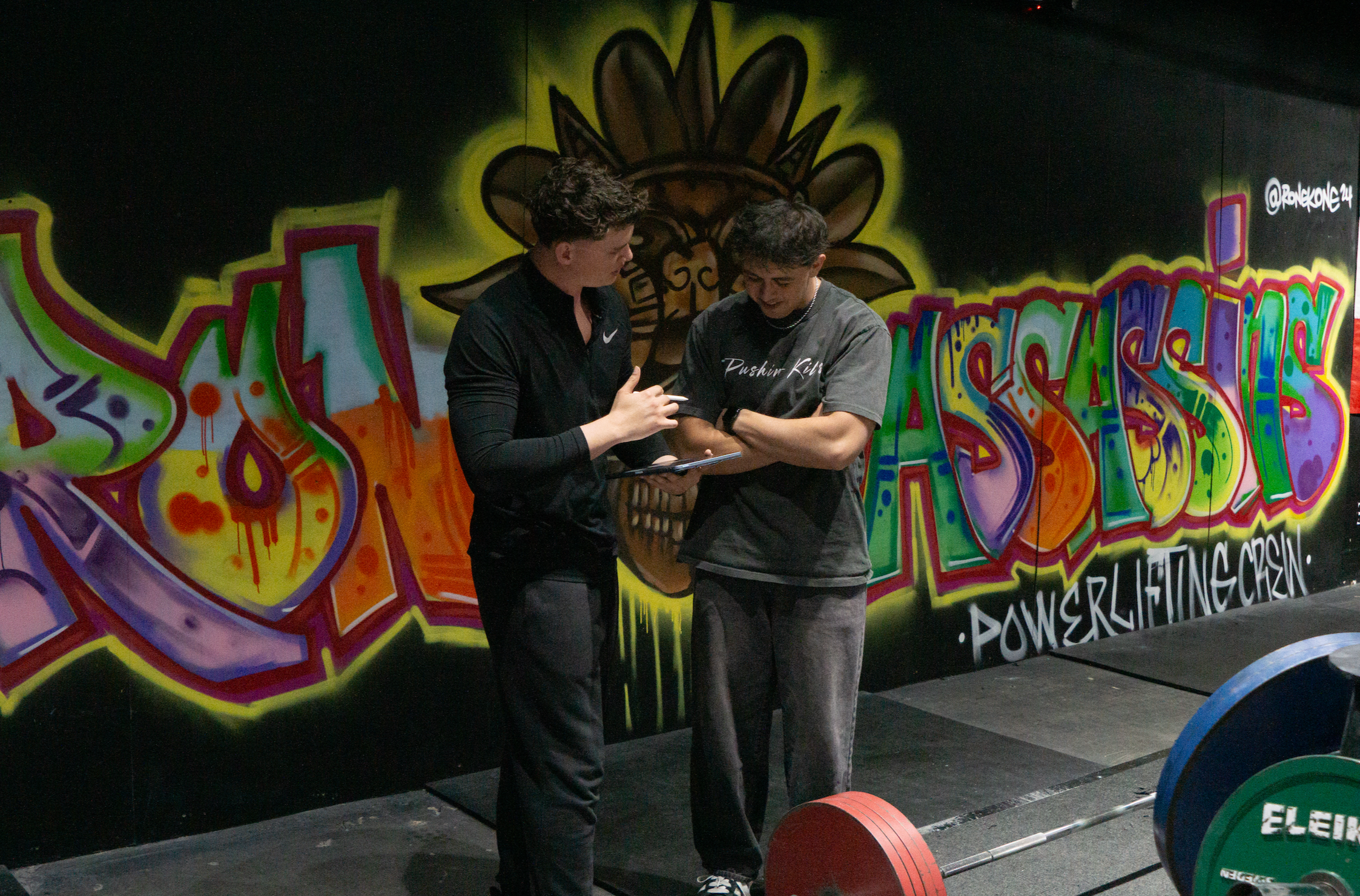 Two young men standing in front of graffiti art on a wall, one holding a clipboard and both looking at it while talking. The background features colorful graffiti with words and abstract designs.