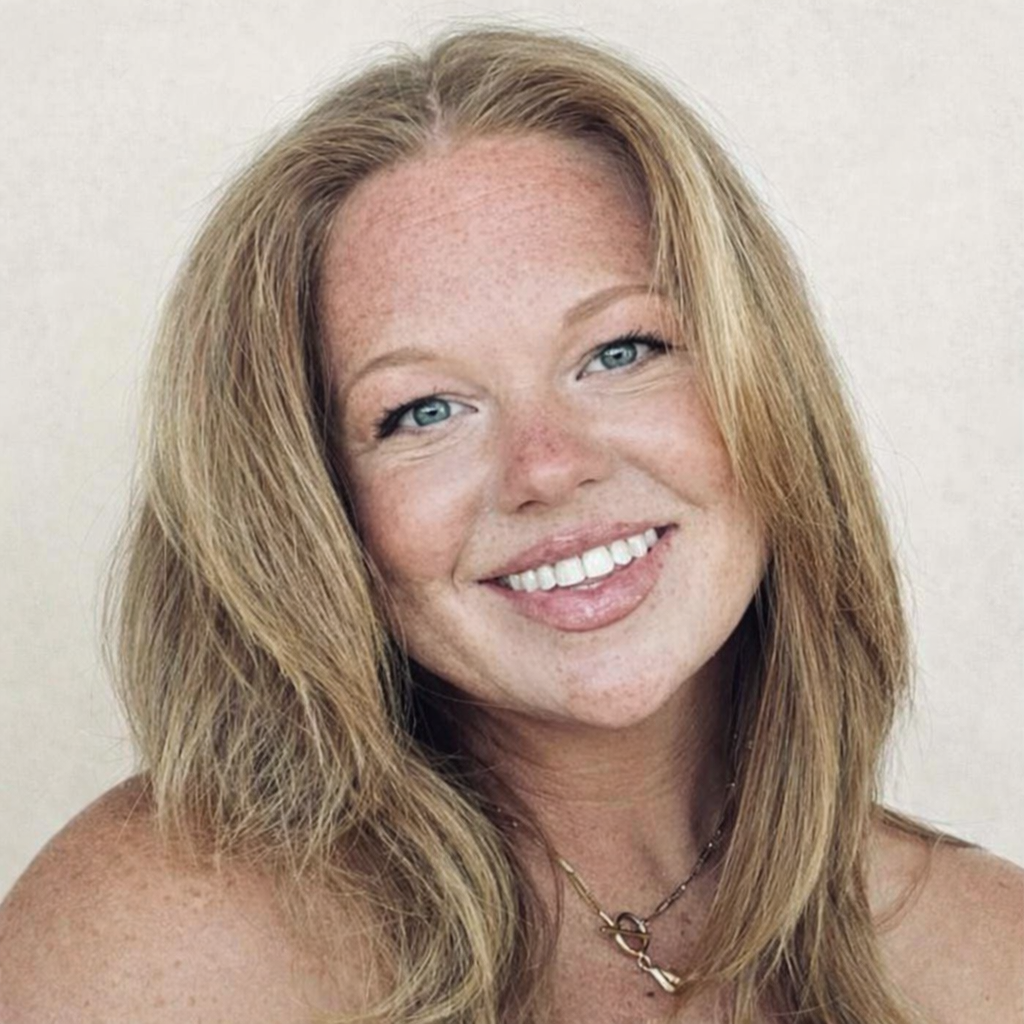 Close-up of a smiling woman with shoulder-length red hair, blue eyes, and freckles, wearing a necklace, against a plain background.