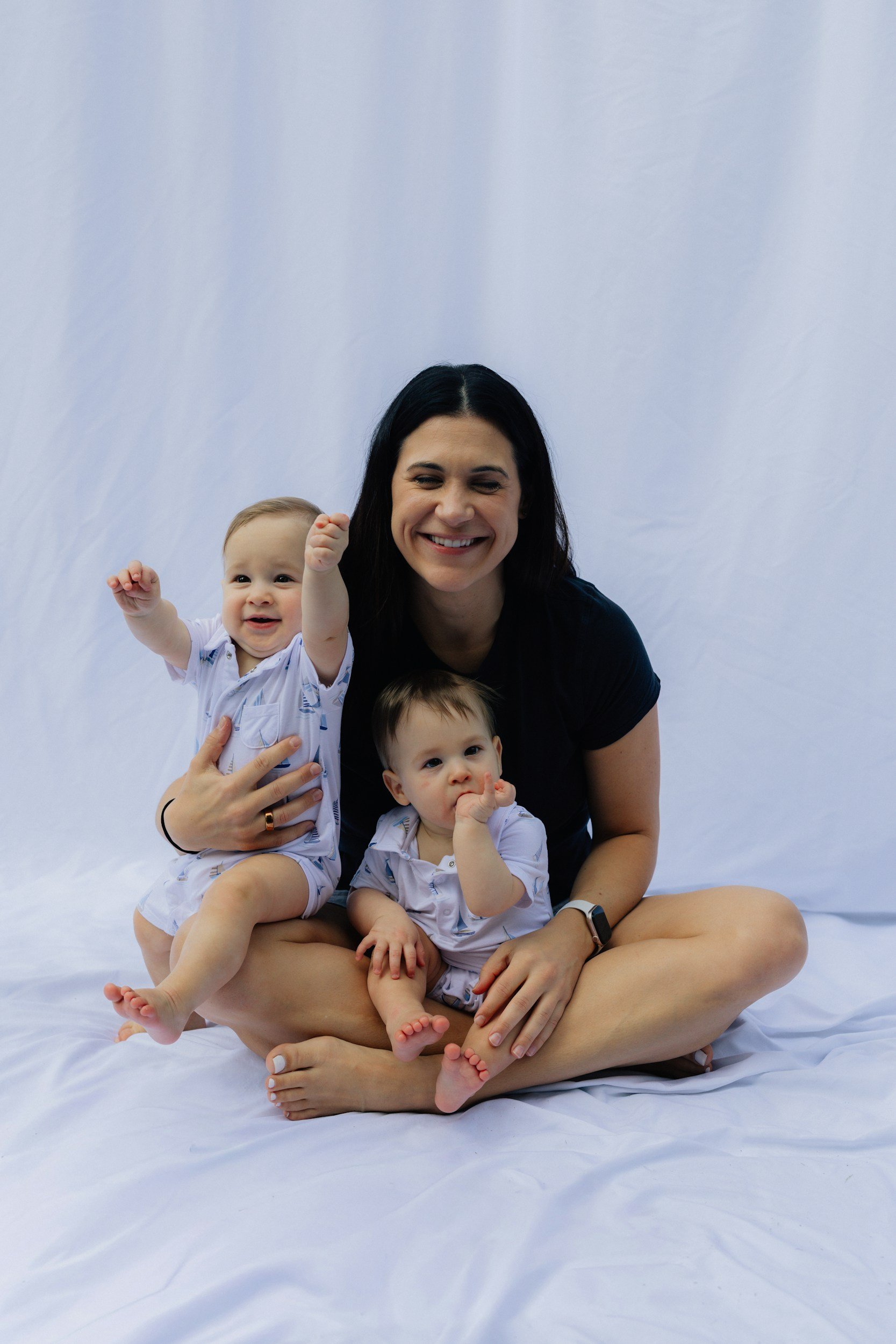 A woman with black hair and a black shirt sitting cross-legged on a white surface, smiling with three babies. The babies are dressed in matching white pajamas with blue designs. Two babies are sitting on her lap, one with light hair and one with darker hair. The third baby, with light hair, is sitting on her crossed legs, holding a pacifier. The background is plain white.