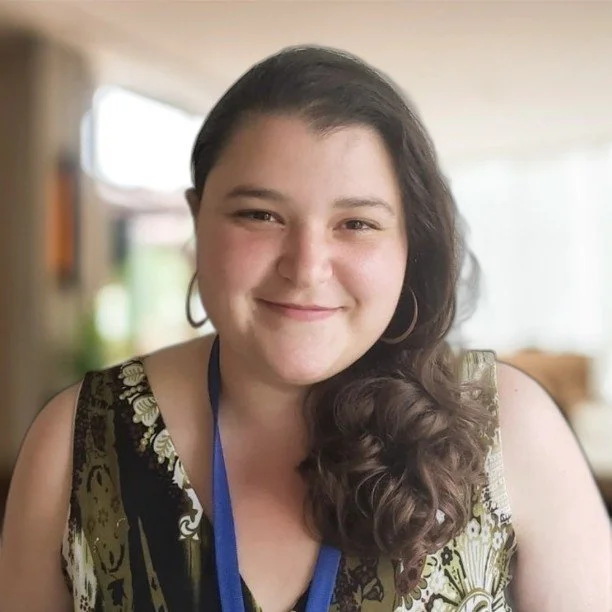 Close-up photo of a smiling woman with dark brown hair and hoop earrings, wearing a patterned sleeveless top and a lanyard, in a bright indoor setting.