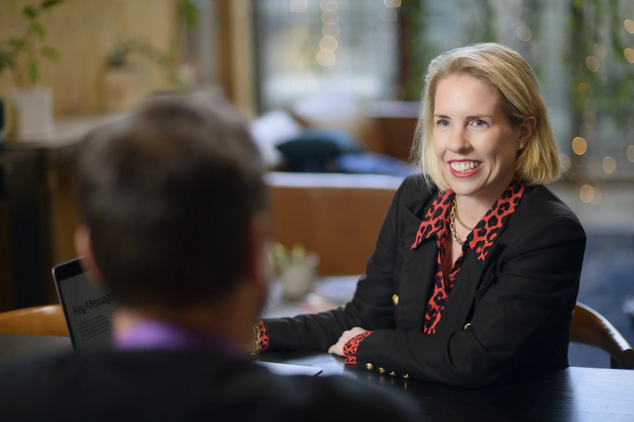 A blonde woman smiling and talking to someone in a cafe or restaurant with greenery and natural light in the background.