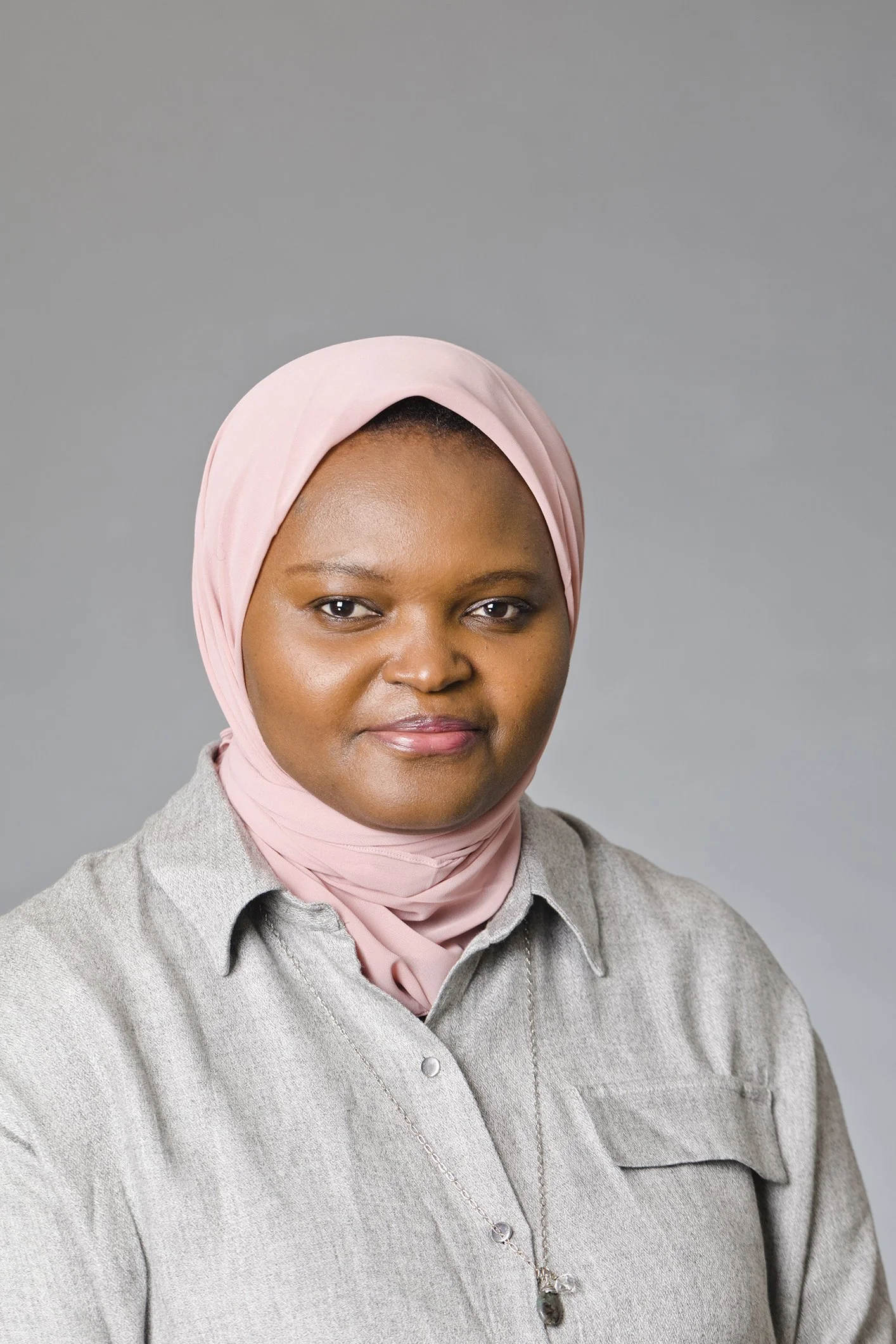 A woman with dark skin and wearing a light pink headscarf, gray button-up shirt, and a necklace with a pendant, standing against a plain gray background.