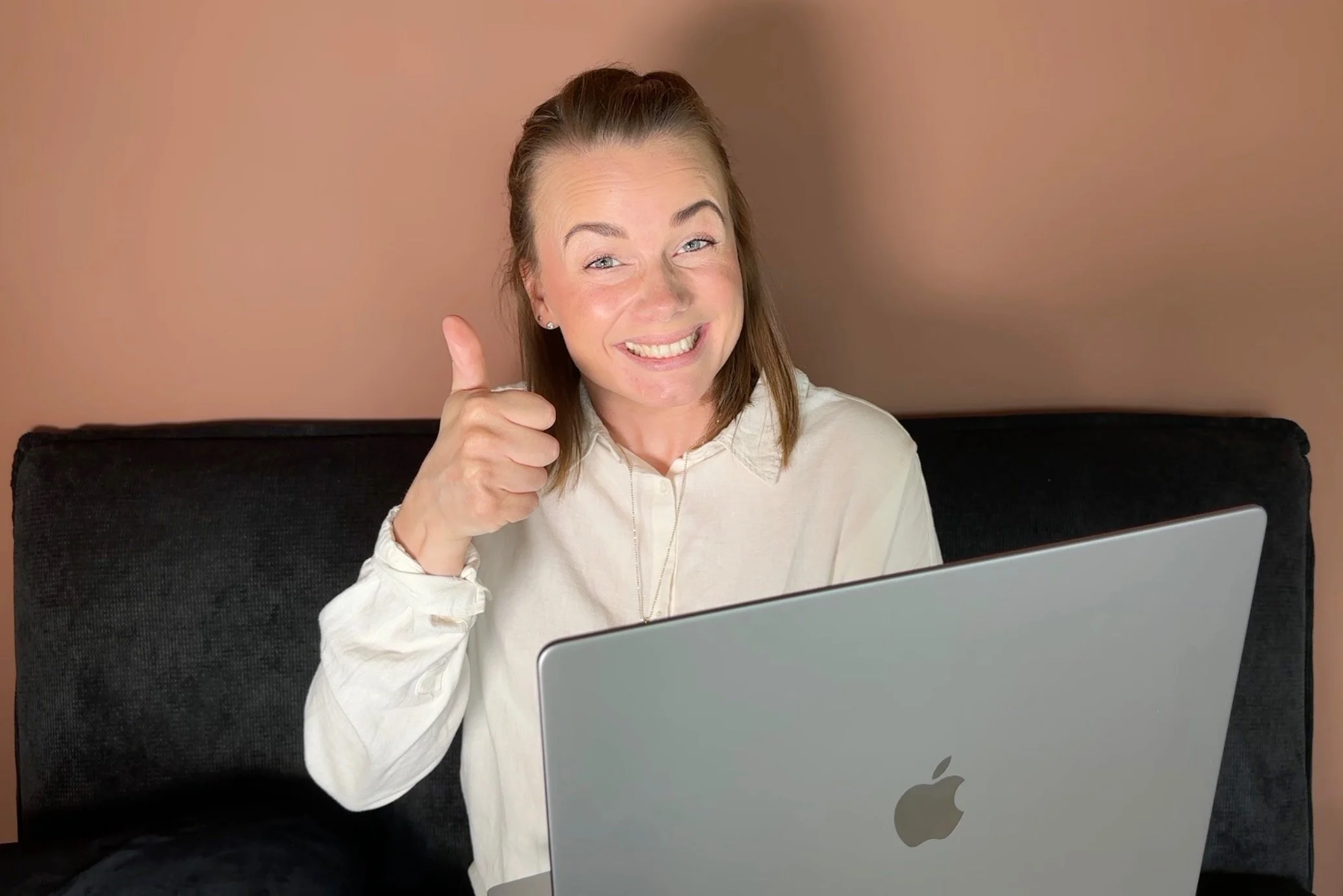 A woman sitting on a black sofa smiling at a laptop with an Apple logo, giving a thumbs-up gesture.