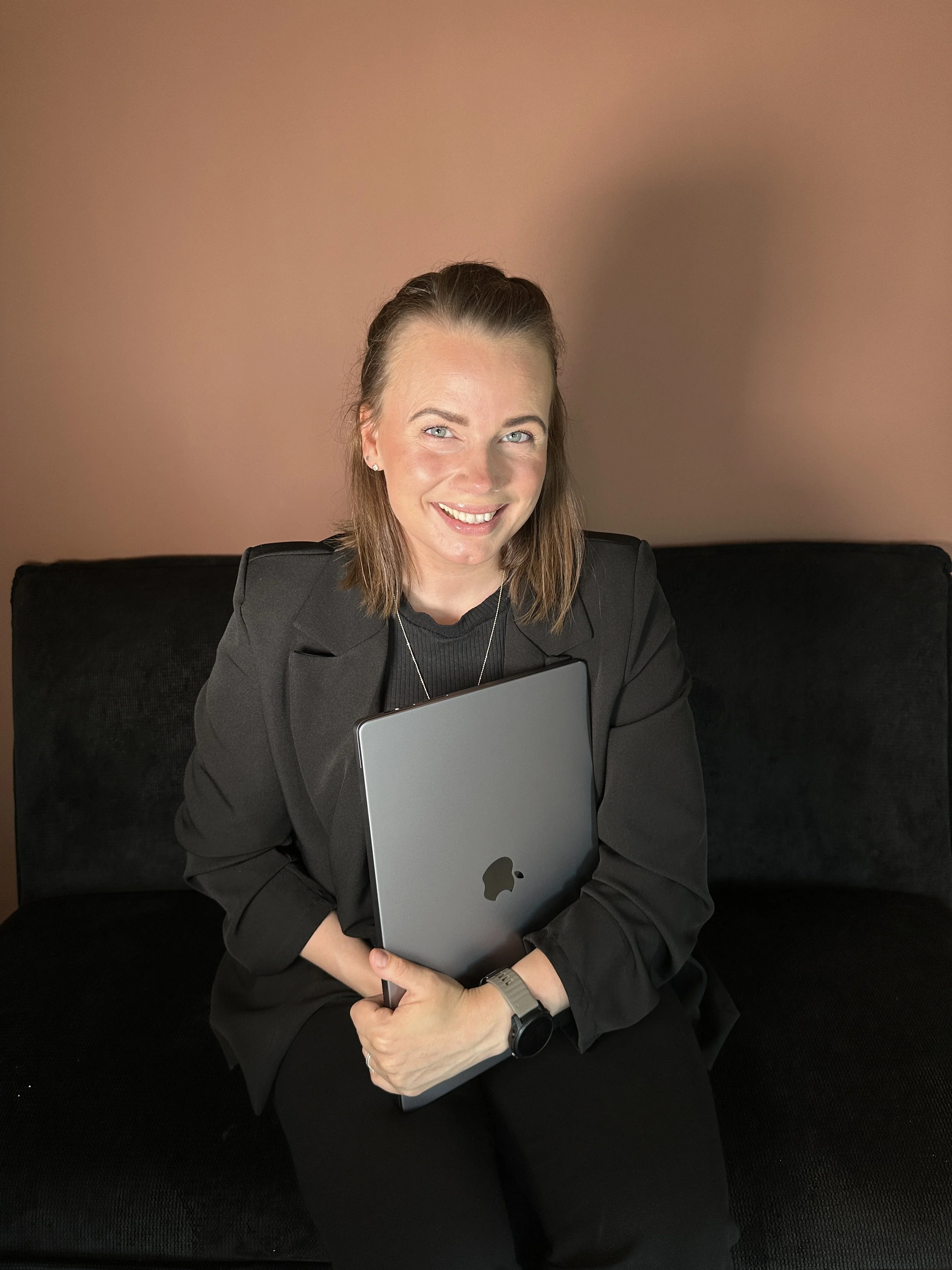 Kvinne sitter med MacBook i sofa og smiler – jobber med nettside og digitale løsninger hjemmefra.