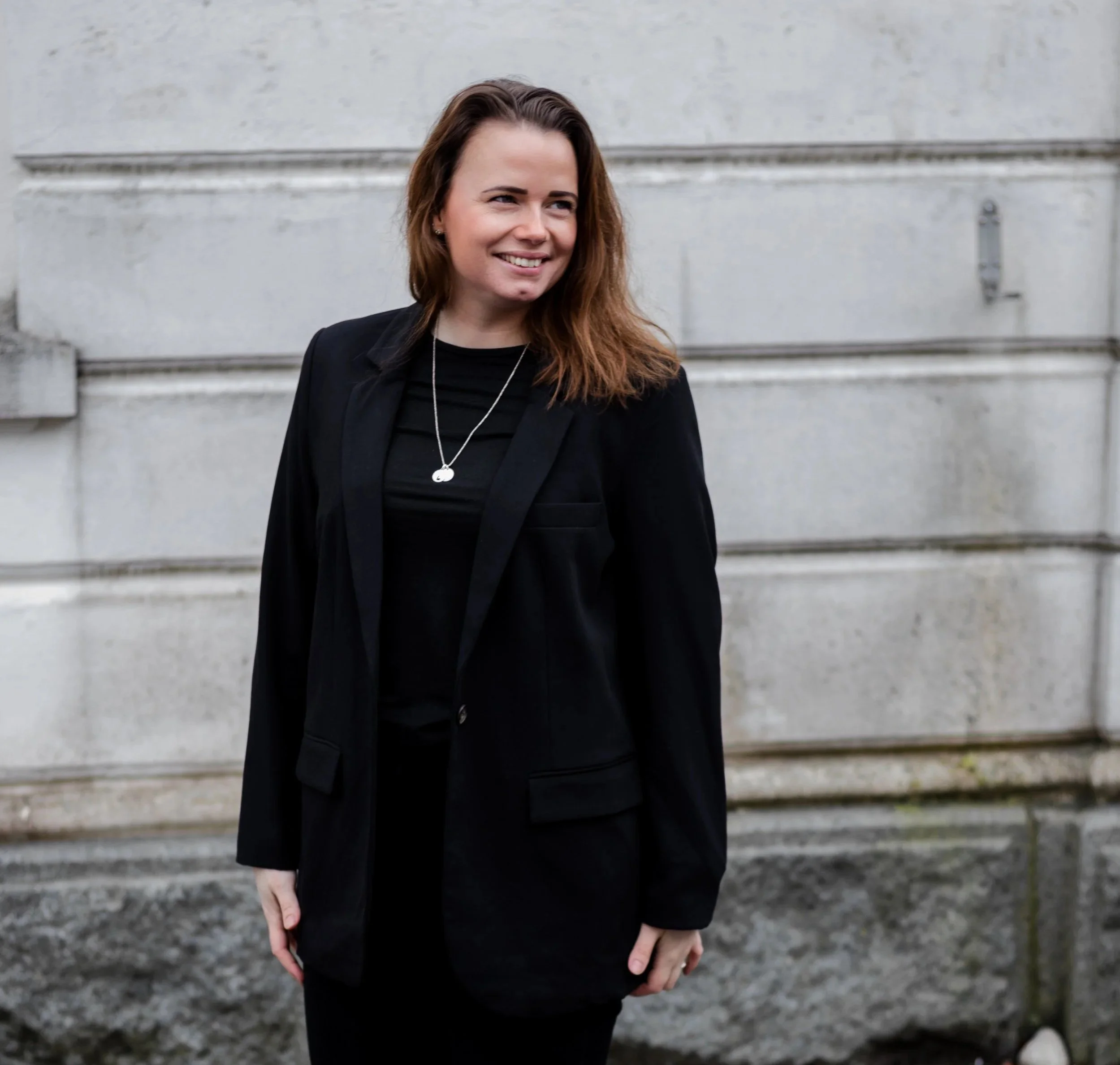 Kvinne i svart blazer smiler utendørs foran steinvegg – bygger personlig merkevare og business.