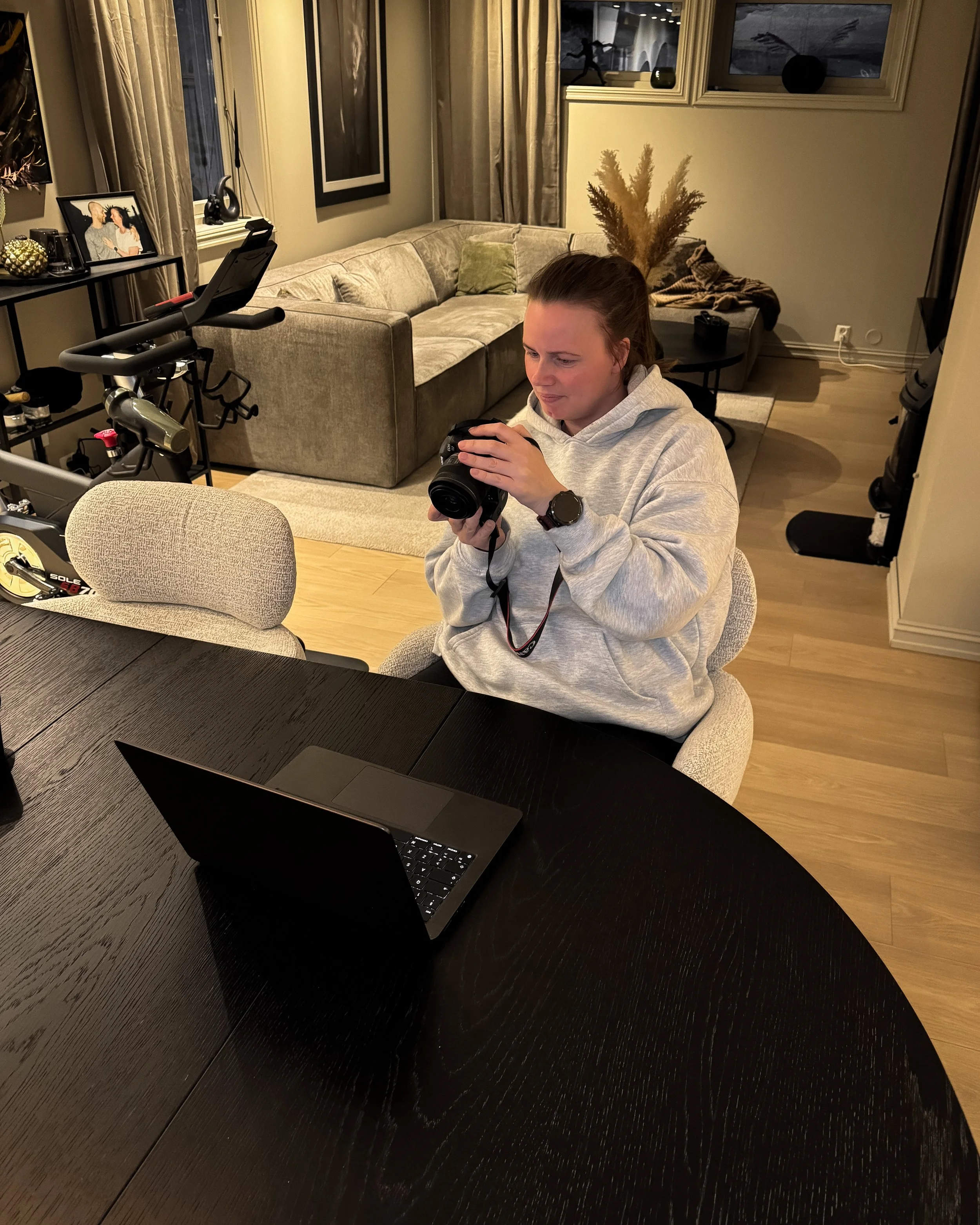 Kvinne i grå hettegenser sitter ved bord med laptop og kamera – jobber med innhold og nettside for egen business.