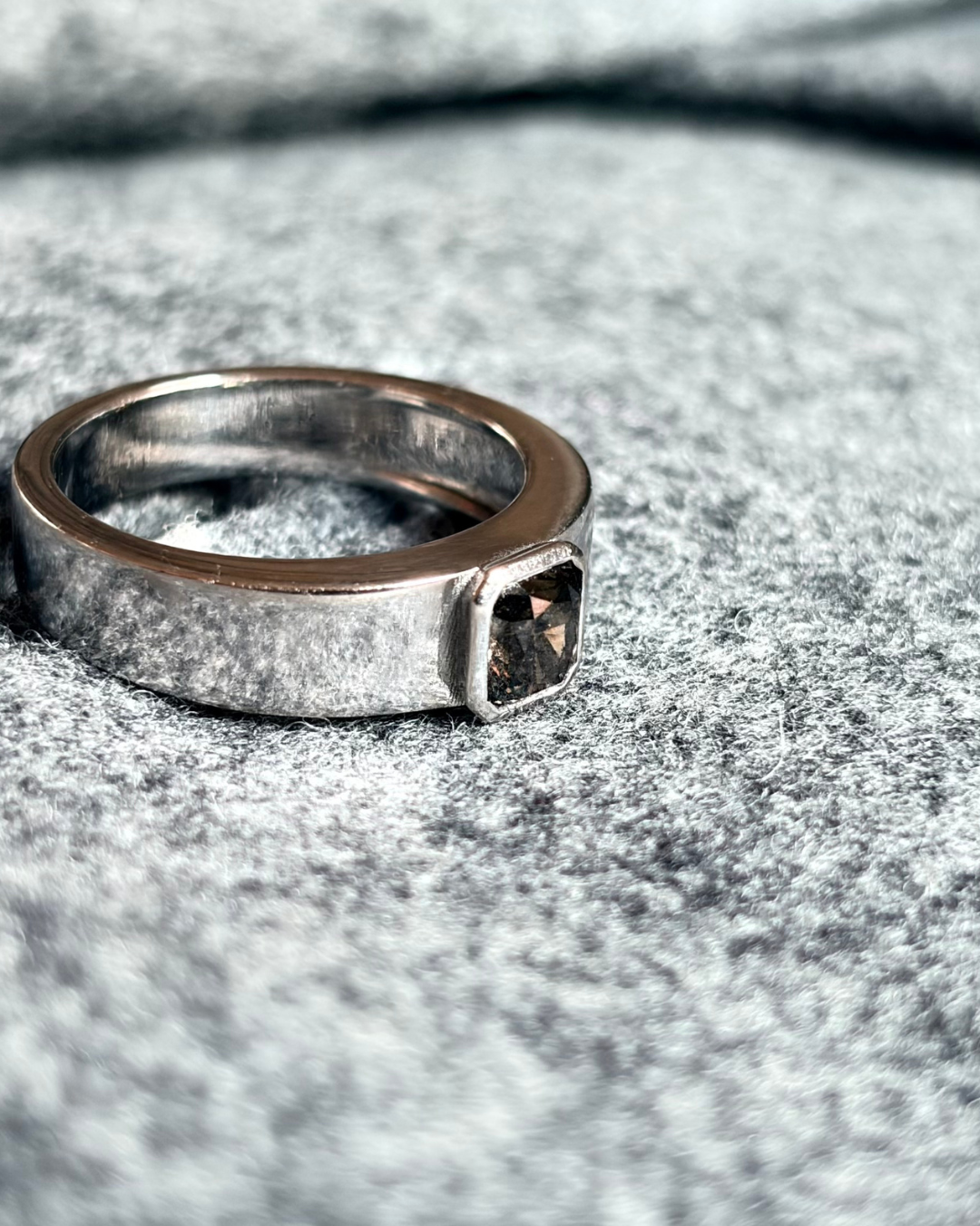 A 18k white gold ring with a fancy colored diamond is placed on a gray fabric surface.