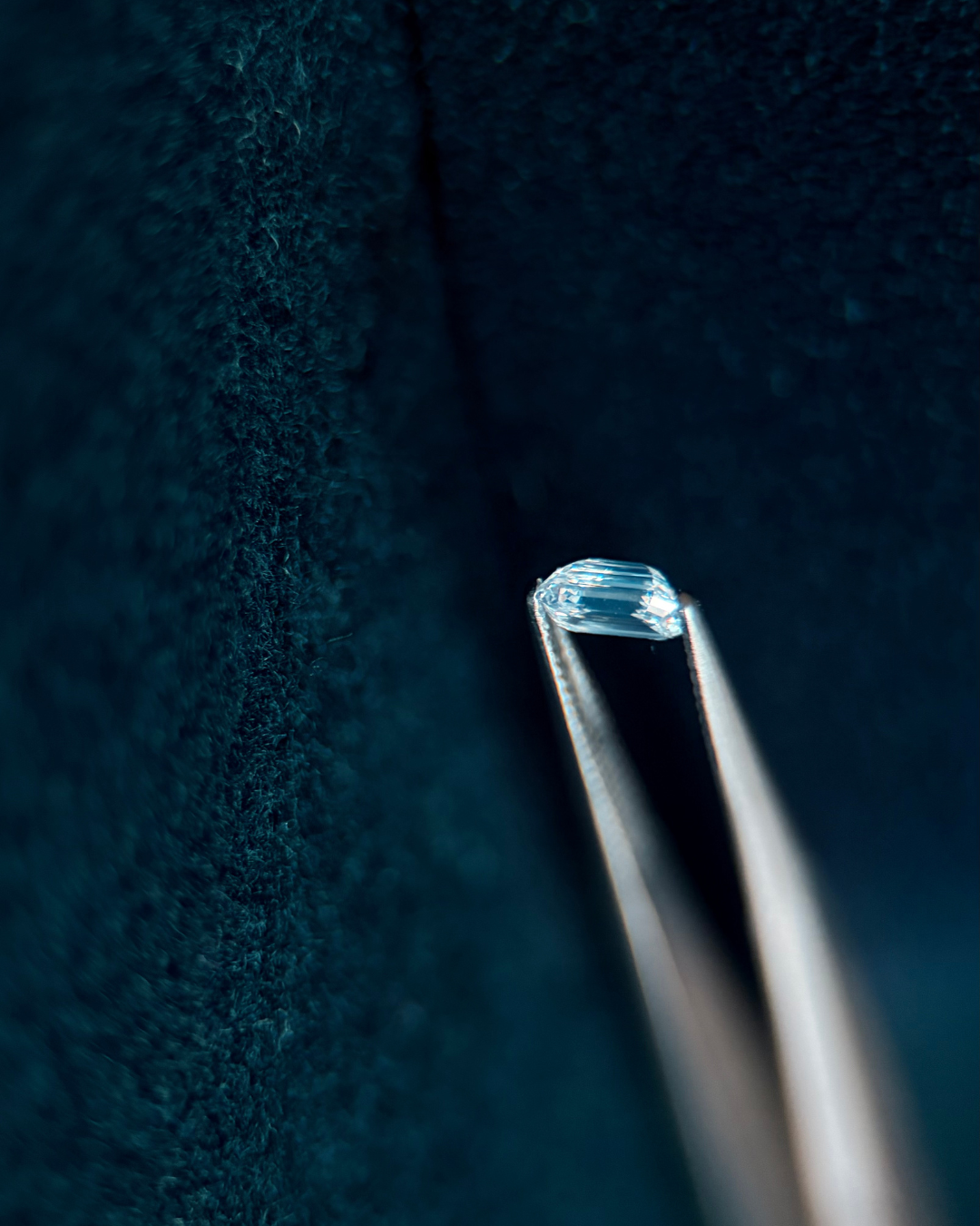 Close-up of a pair of metal tweezers holding a transparent, faceted gemstone against a dark textured background.