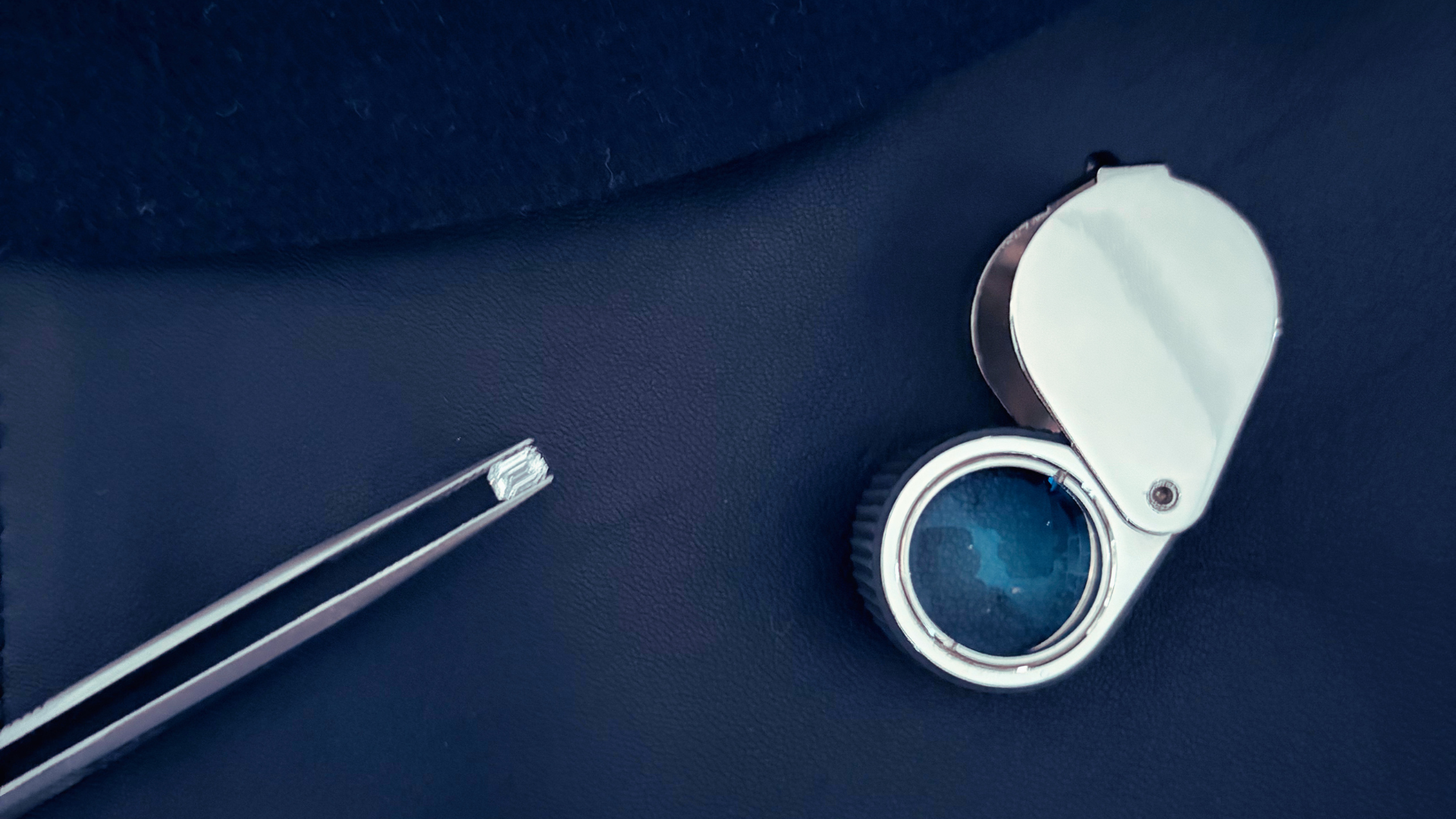 A gemologist tweezer and a loupe dark blue textured surface, with a pair of metal tweezers nearby.