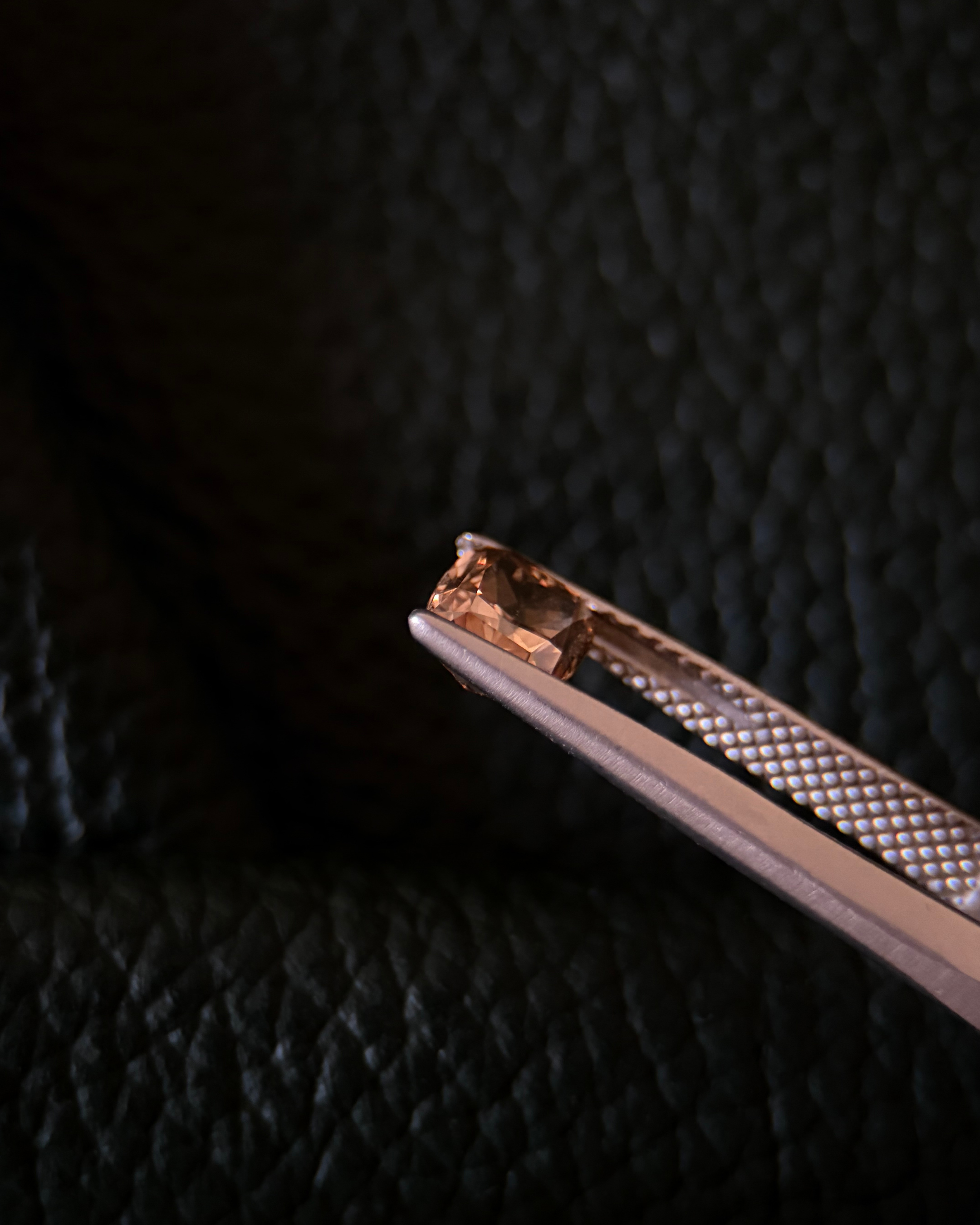 Close-up of a orange-yellow diamond held with metal tweezers against a dark blue leather background.