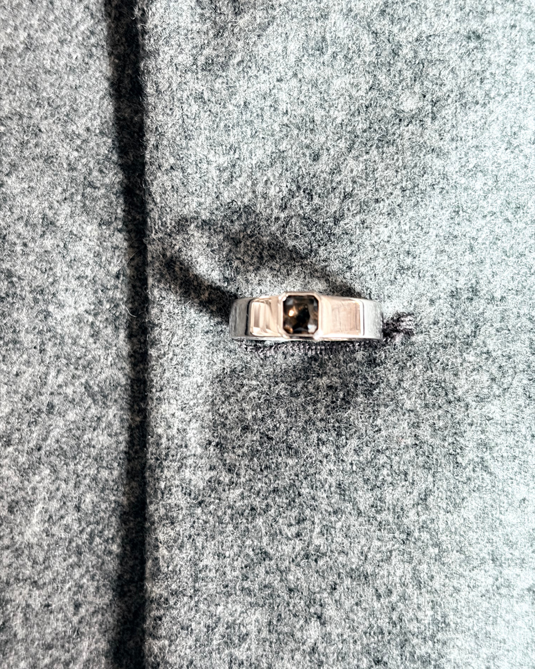 A silver ring with a rectangular dark gemstone on a gray textured fabric surface.