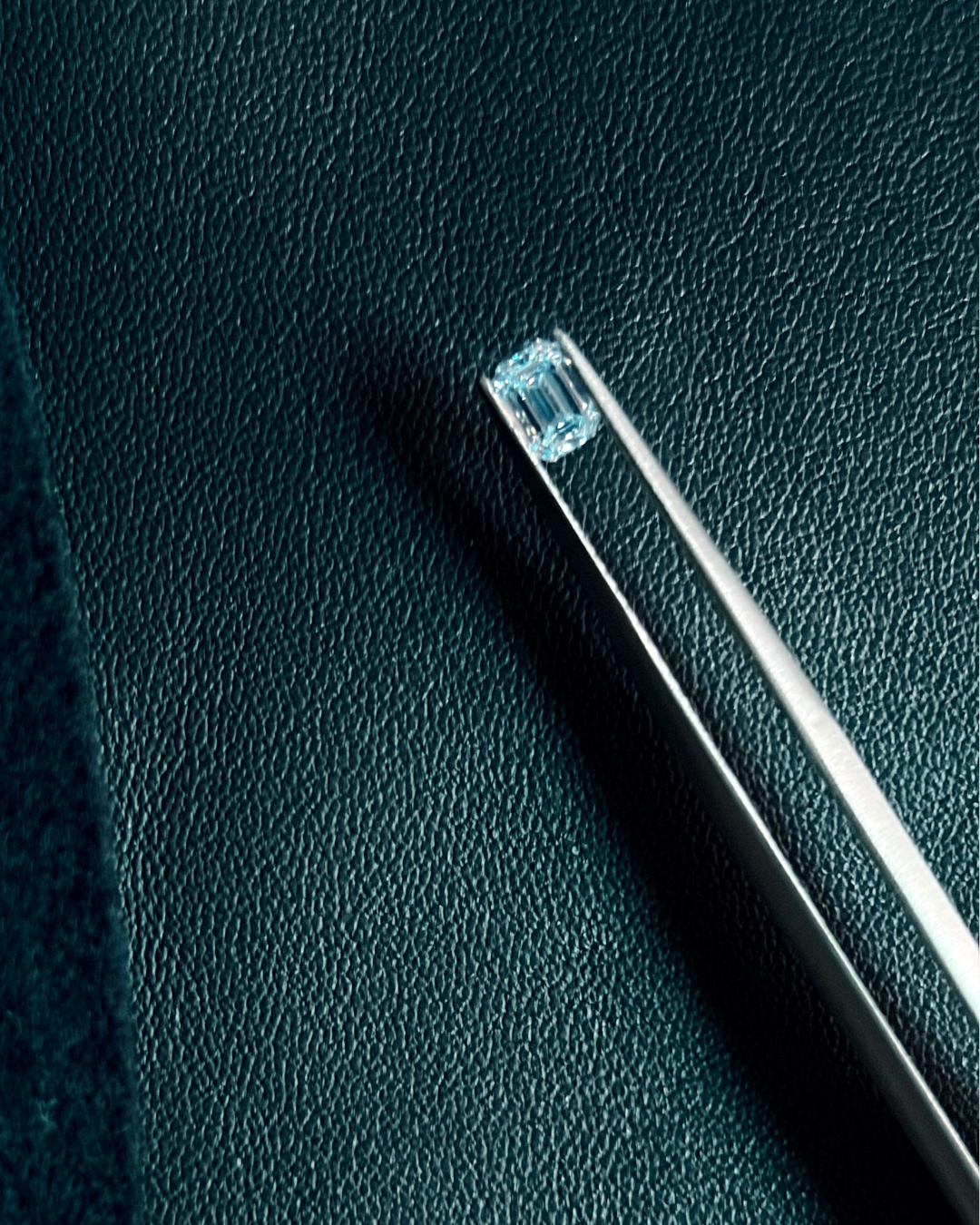 A silver pair of tweezers holding a rectangular emerald-cut blue gemstone above a textured dark green surface.