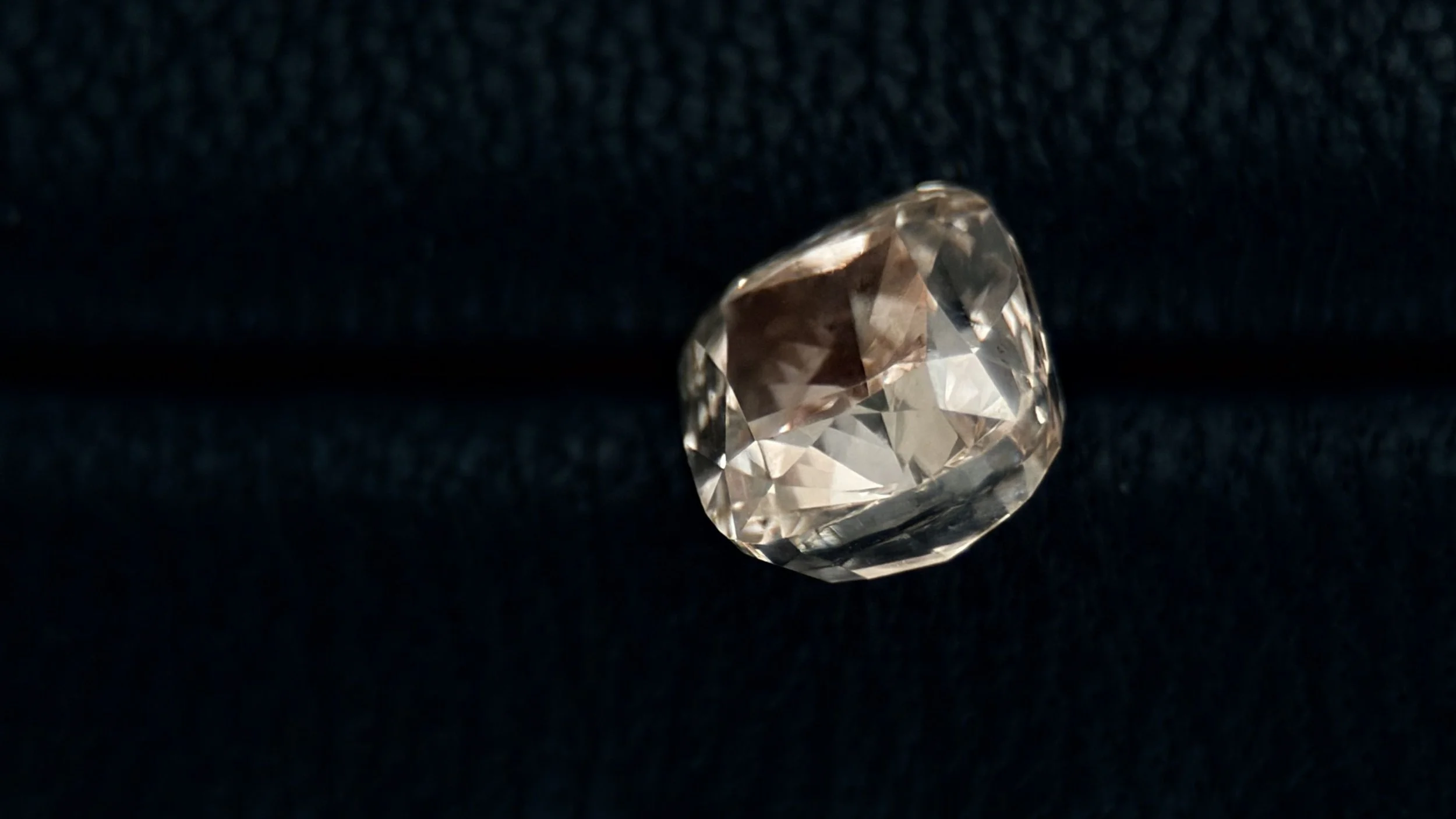 A close-up of a clear, faceted gemstone or diamond resting on a dark, textured surface.