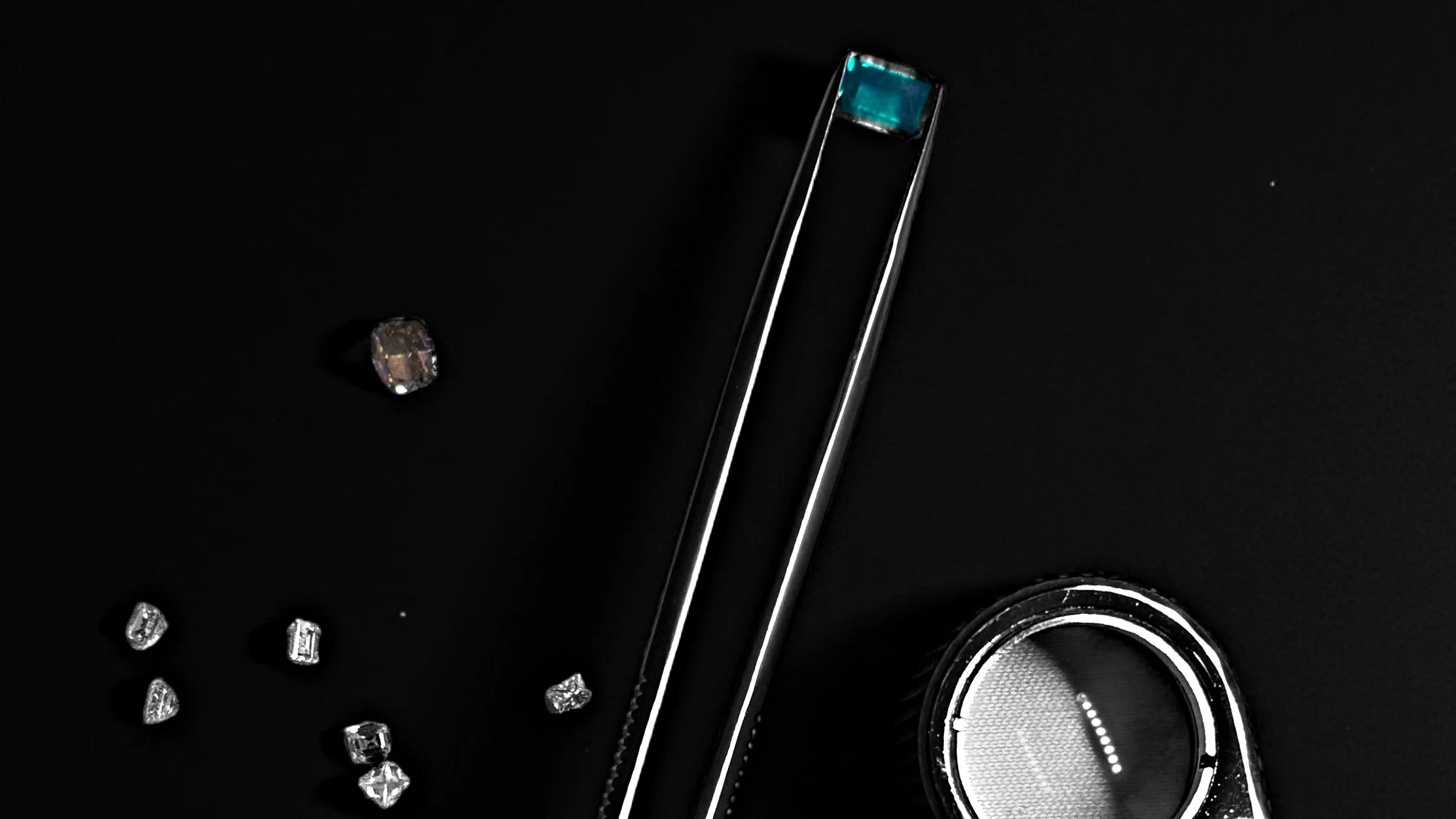 An emerald held in gemological tweezers beside a selection of natural diamonds and a loupe
