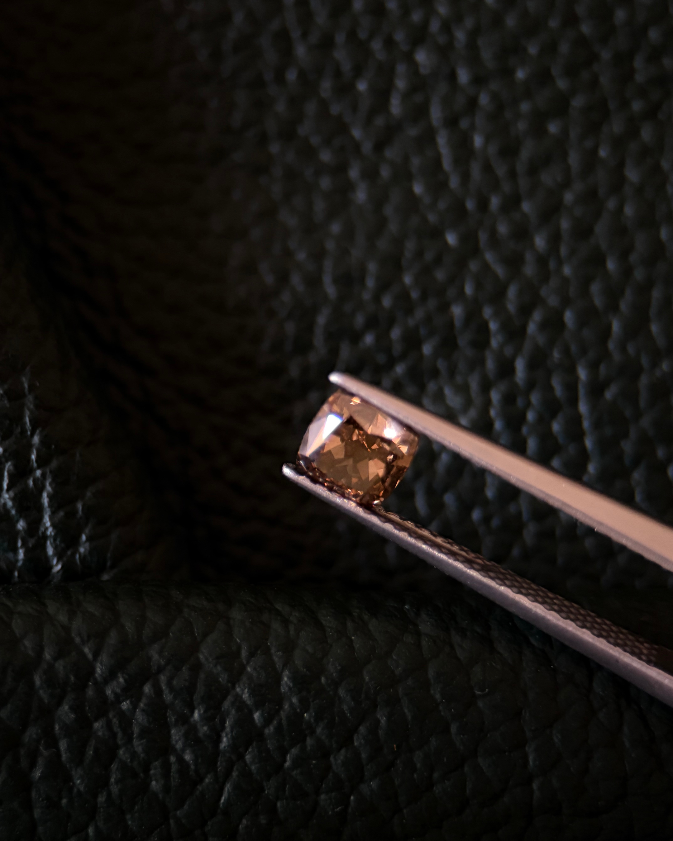 A small, square-shaped, brown gemstone held with metal tweezers against a dark, textured background.