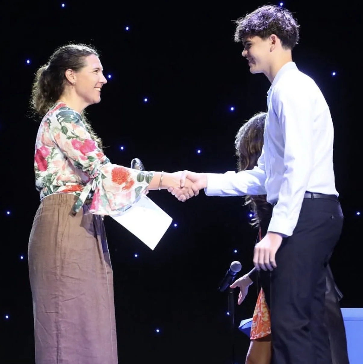 A woman and a young man shake hands on stage during an event, with both smiling and looking at each other, against a background decorated with small, glowing lights.