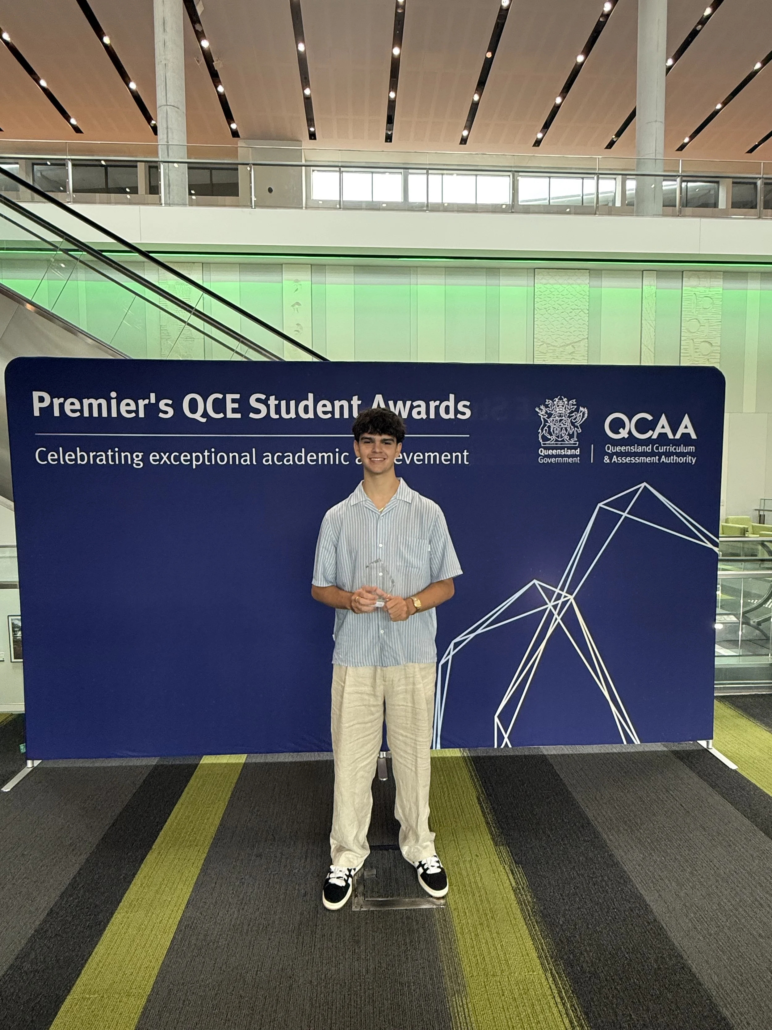 Young man holding an award standing in front of a blue backdrop at the Premier's QCE Student Awards, organized by the Queensland Curriculum & Assessment Authority.