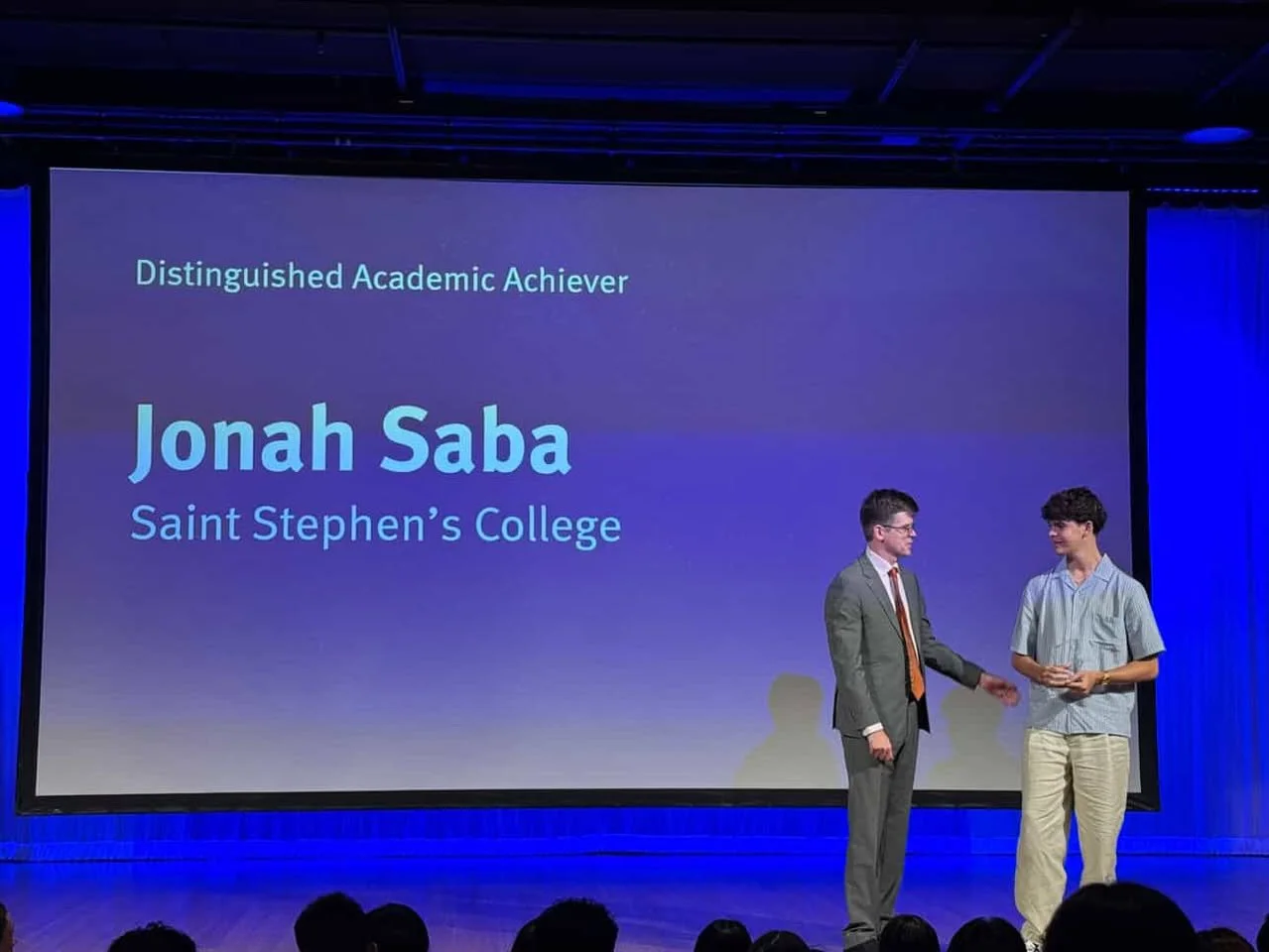 Two young men on stage during an award ceremony, one presenting an award to the other, with a large screen behind them displaying the text 'Distinguished Academic Achiever Jonah Saba Saint Stephen's College'.