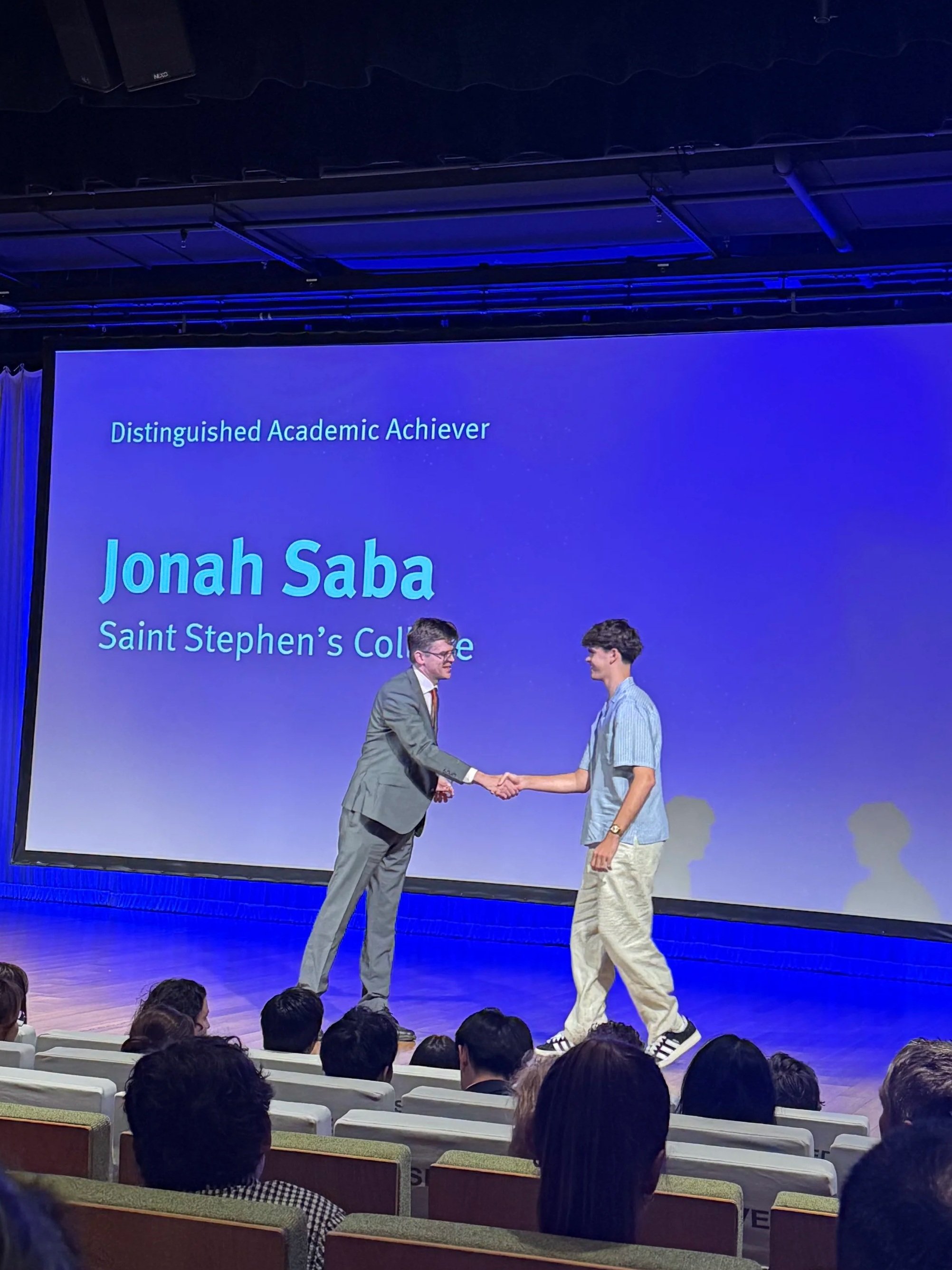 A graduation or award ceremony with a young man in casual attire receiving an award or recognition from a man in a suit on stage. The background screen displays text about a distinguished academic achiever named Jonah Saba from Saint Stephen’s College.