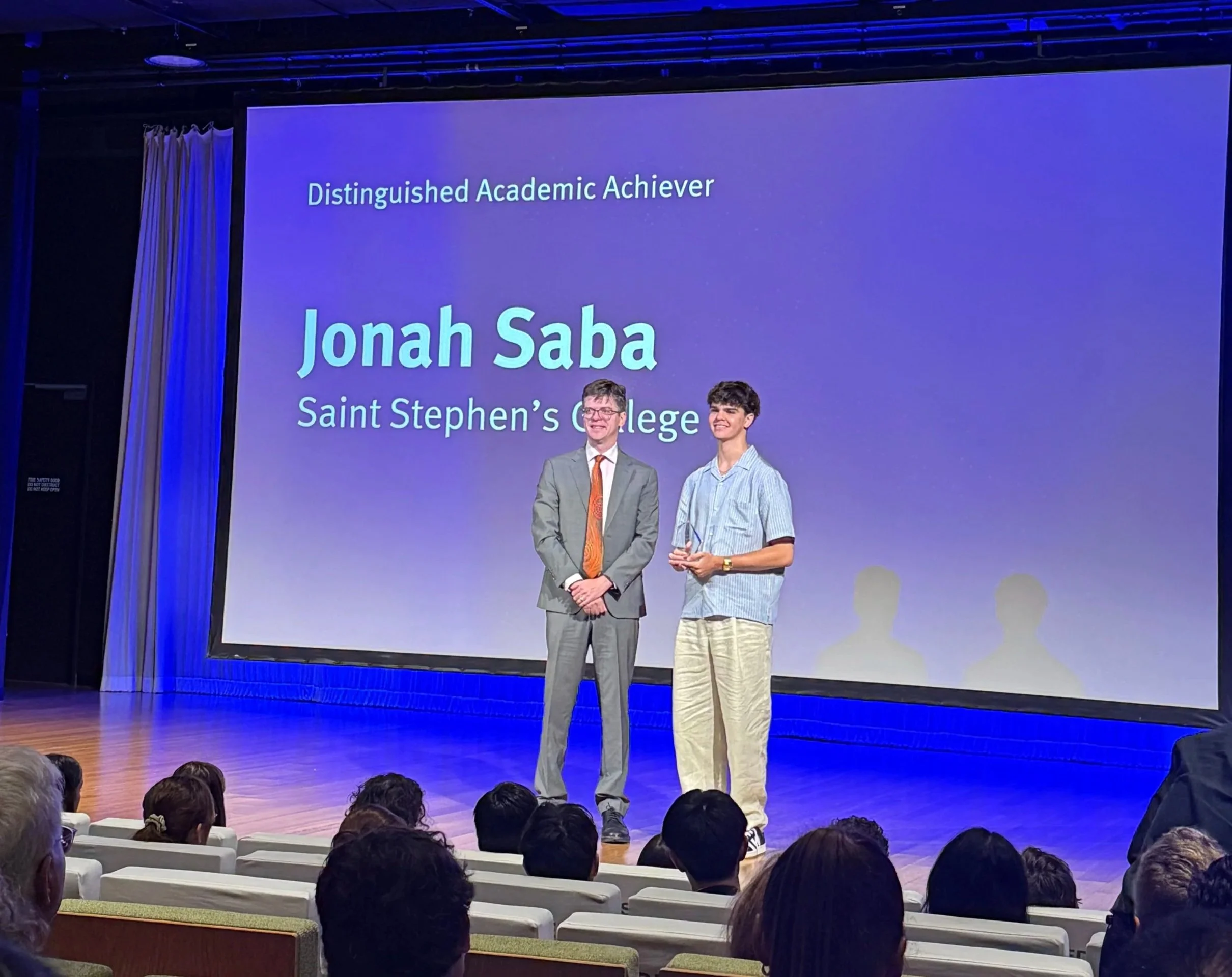 A young man receiving an award on stage at Saint Stephen's College, standing next to a man in a suit. A large screen behind them displays the name 'Jonah Saba' and titles, with an audience seated in front.